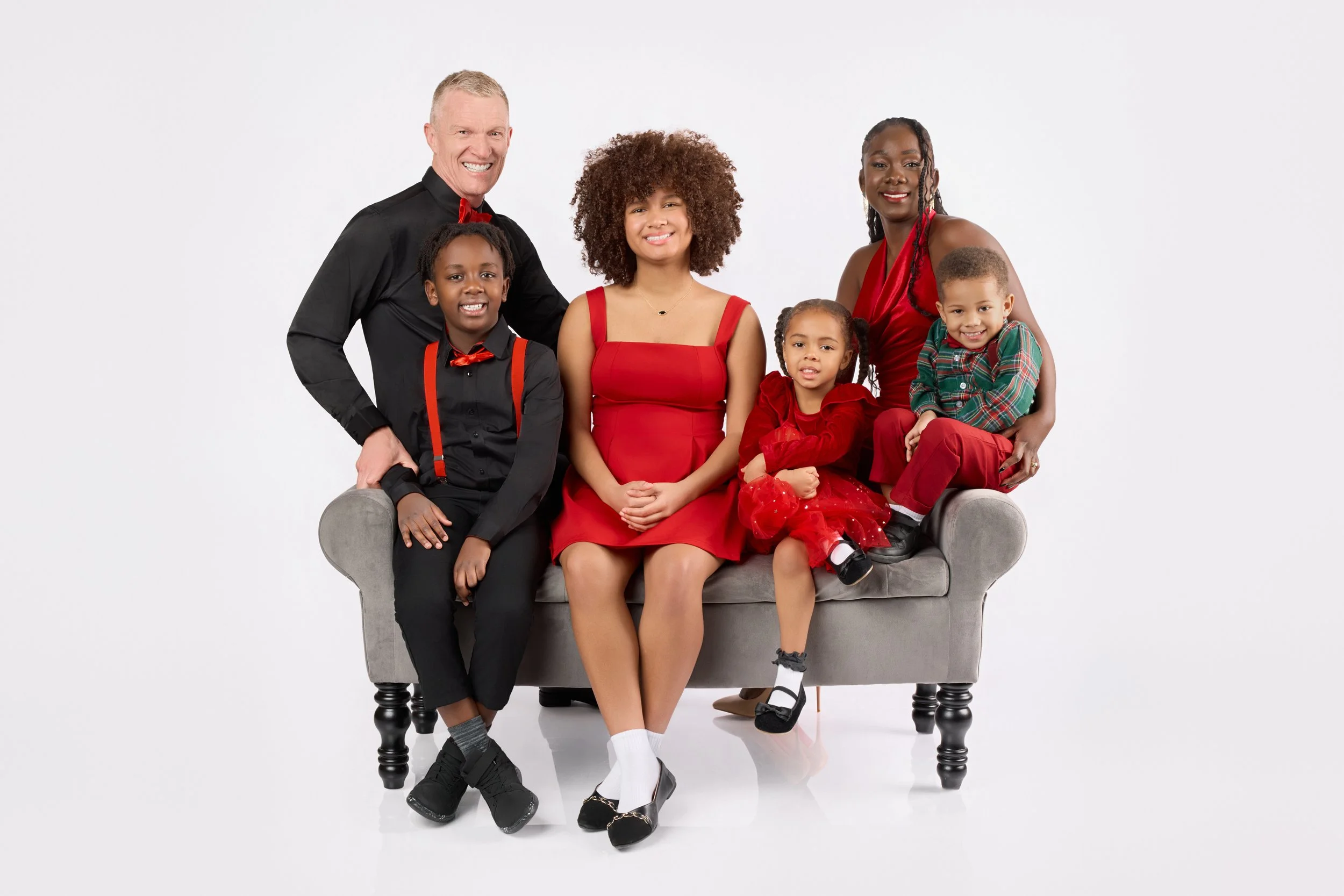 Family of six posing together on a gray sofa, dressed in red and black. Two adults and four children smiling at the camera.