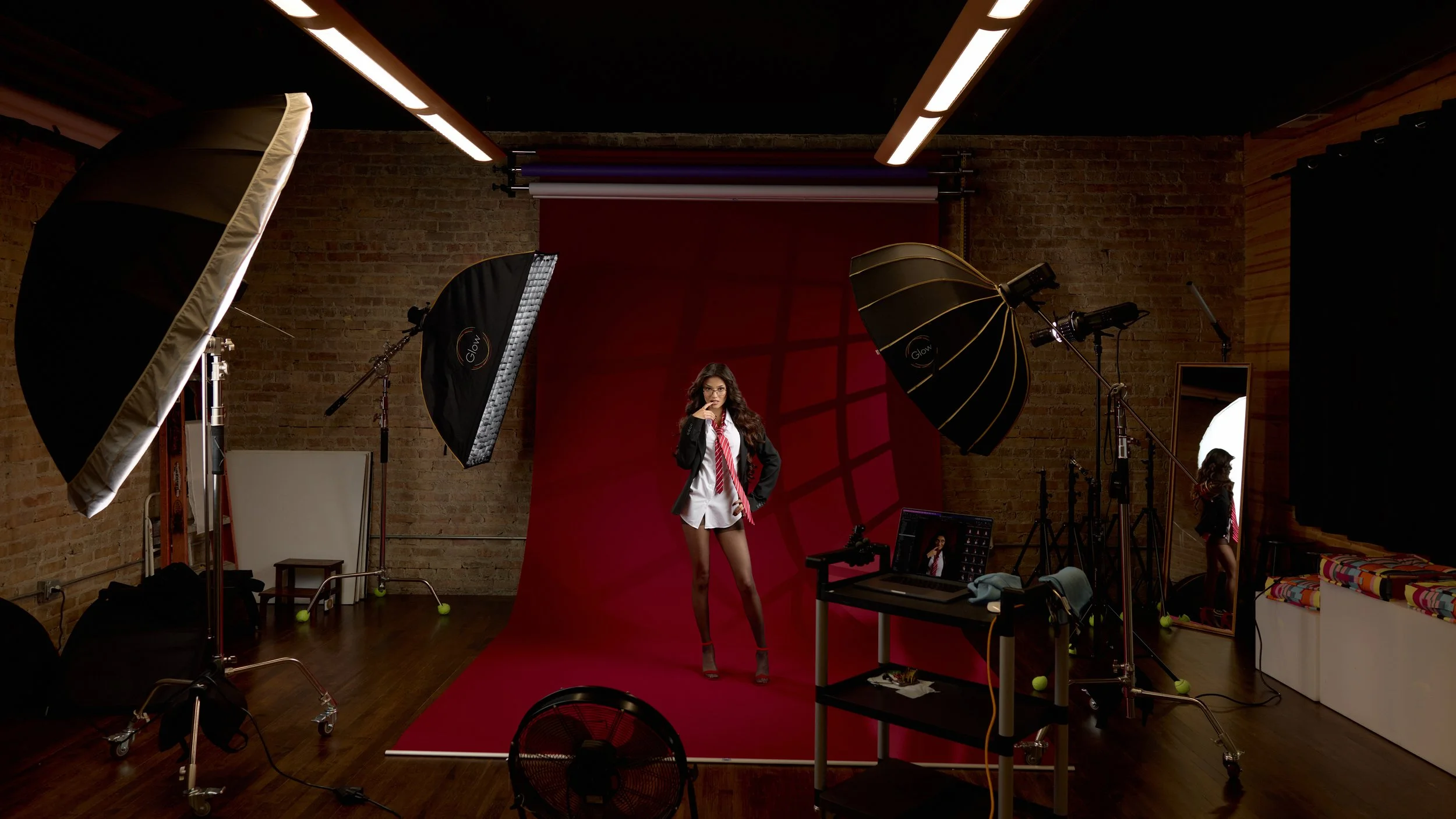Behind the scenes portrait of a studio fashion session with a woman in a schoolgirl outfit standing in front of a red backdrop, surrounded by professional lighting and camera equipment, and a mirror reflecting her.