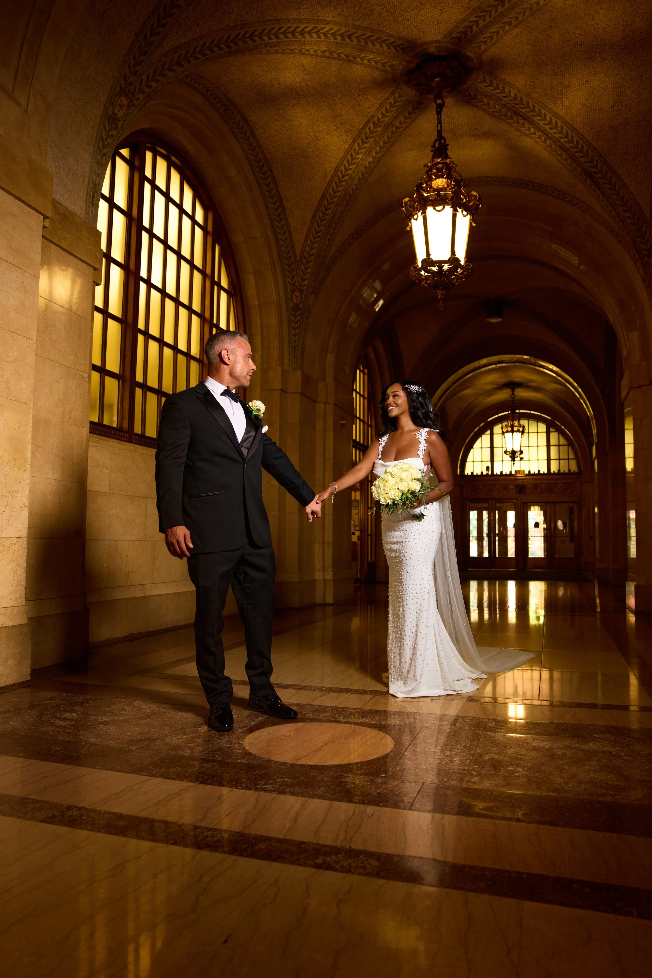 Civil Courthouse wedding of a bride and groom holding hands in a grand, historic building with tall arched windows and warm lighting, celebrating their wedding.
