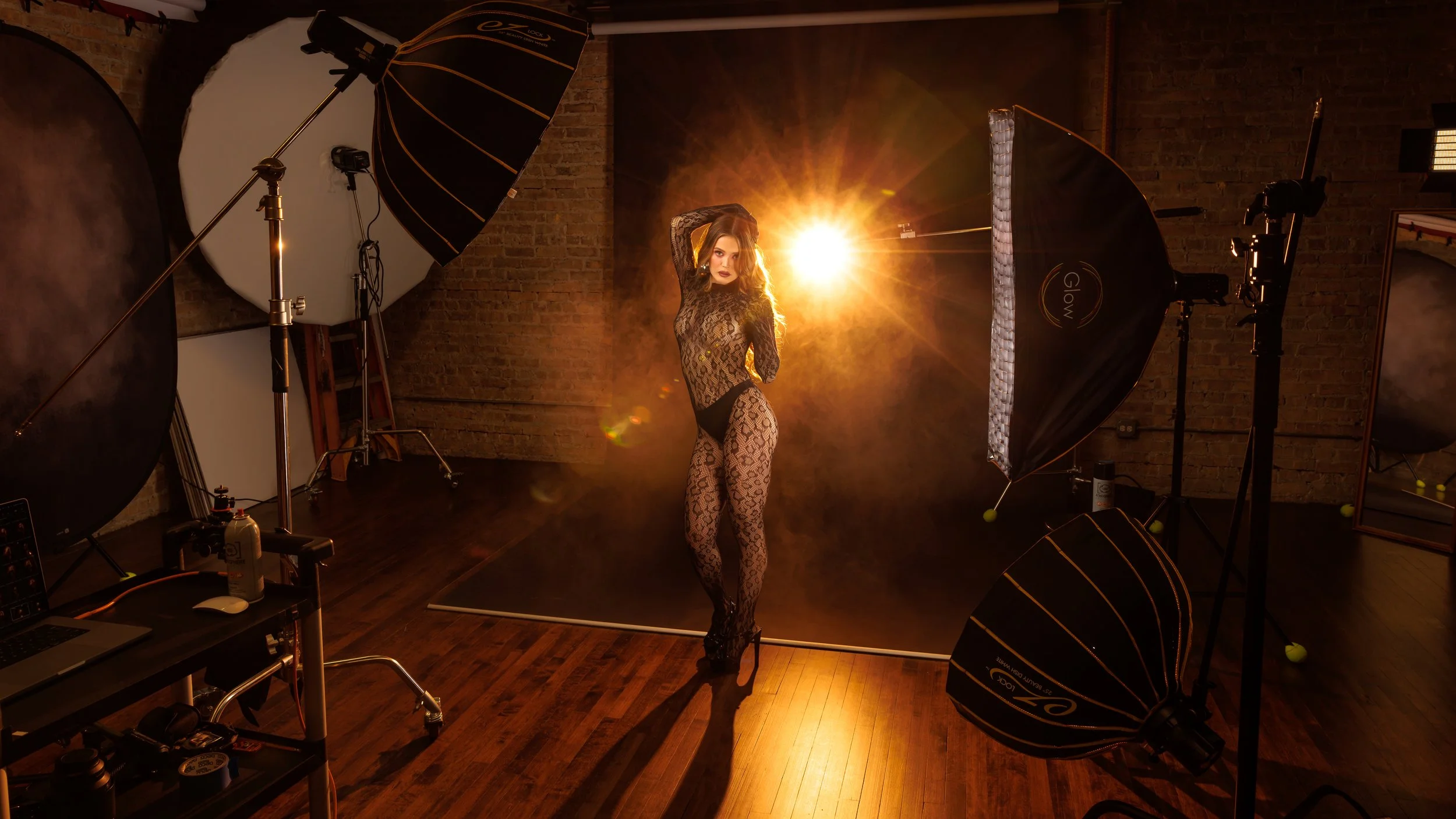 A woman in lace bodysuit and high heels poses in a photography studio with professional lighting and backdrop, illuminated by a bright yellow-golden spotlight.