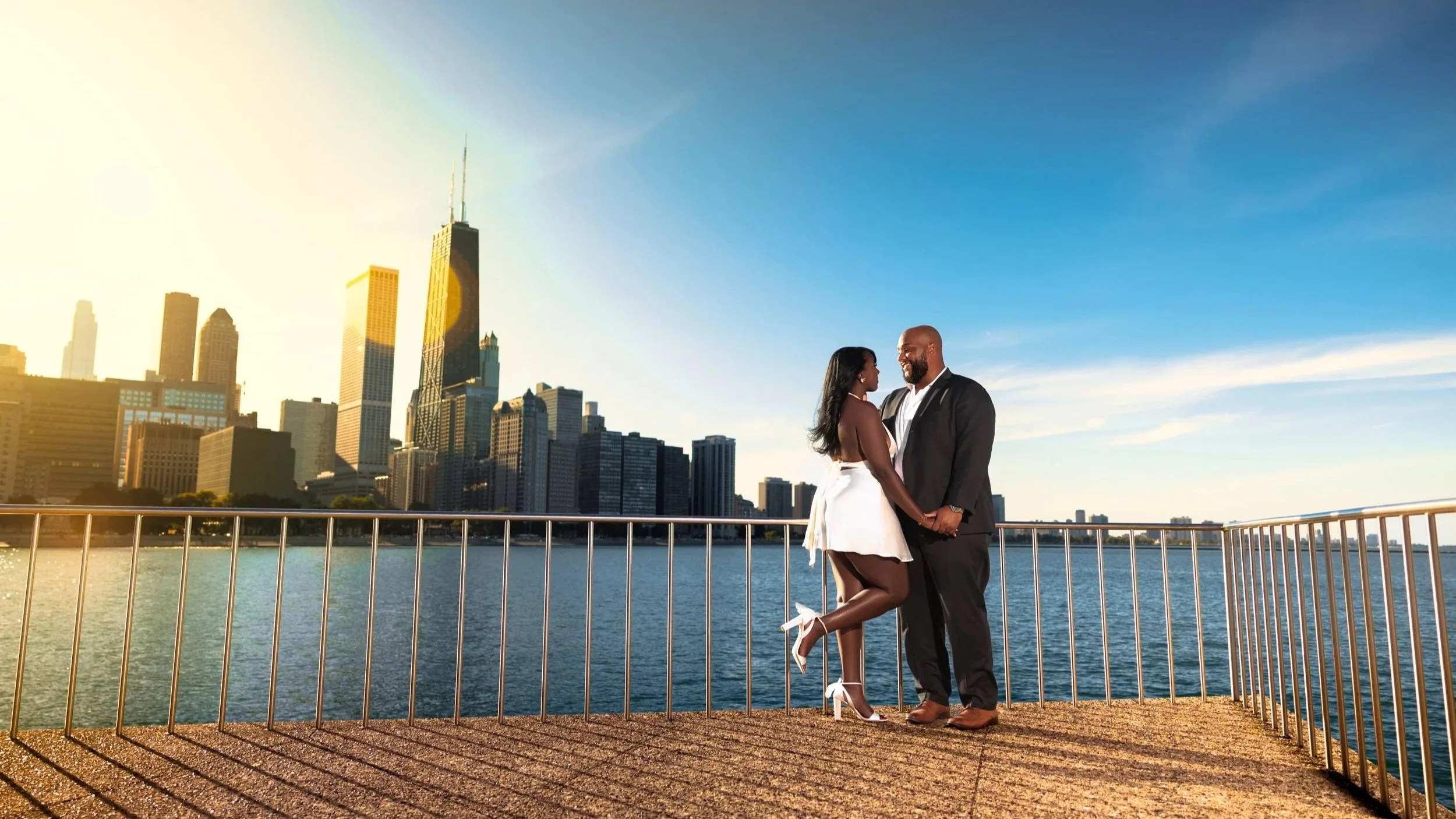 A couple is standing on a waterfront promenade in front of the city skyline at sunset, holding hands and smiling at each other.