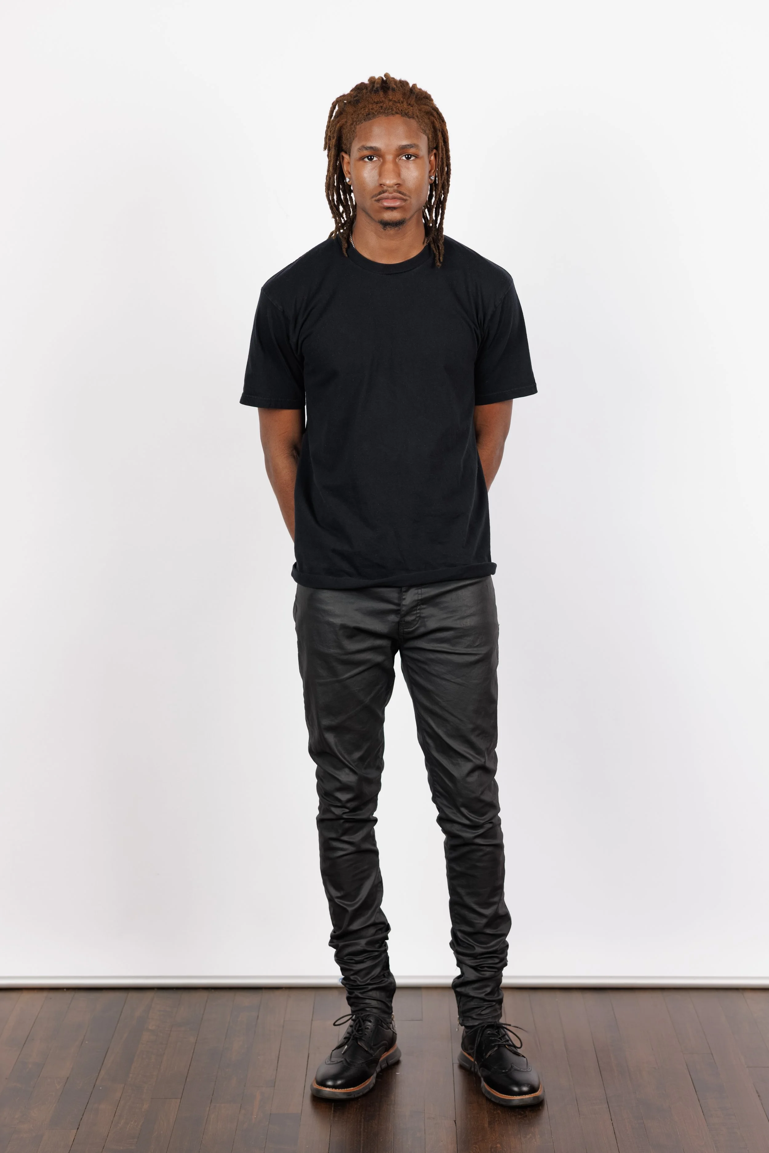 Chicago Modelling Digital of a young man with dreadlocks stands against a plain white background. He wears a black t-shirt, black pants, and black shoes, with his hands behind his back and a serious expression.