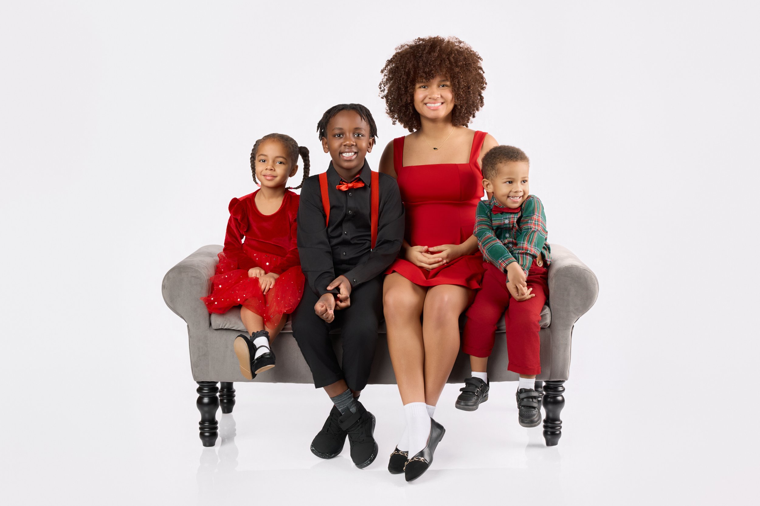 A woman and three children sitting on a gray sofa against a plain white background. The woman and children are dressed in festive red, black, and green holiday attire, smiling at the camera.