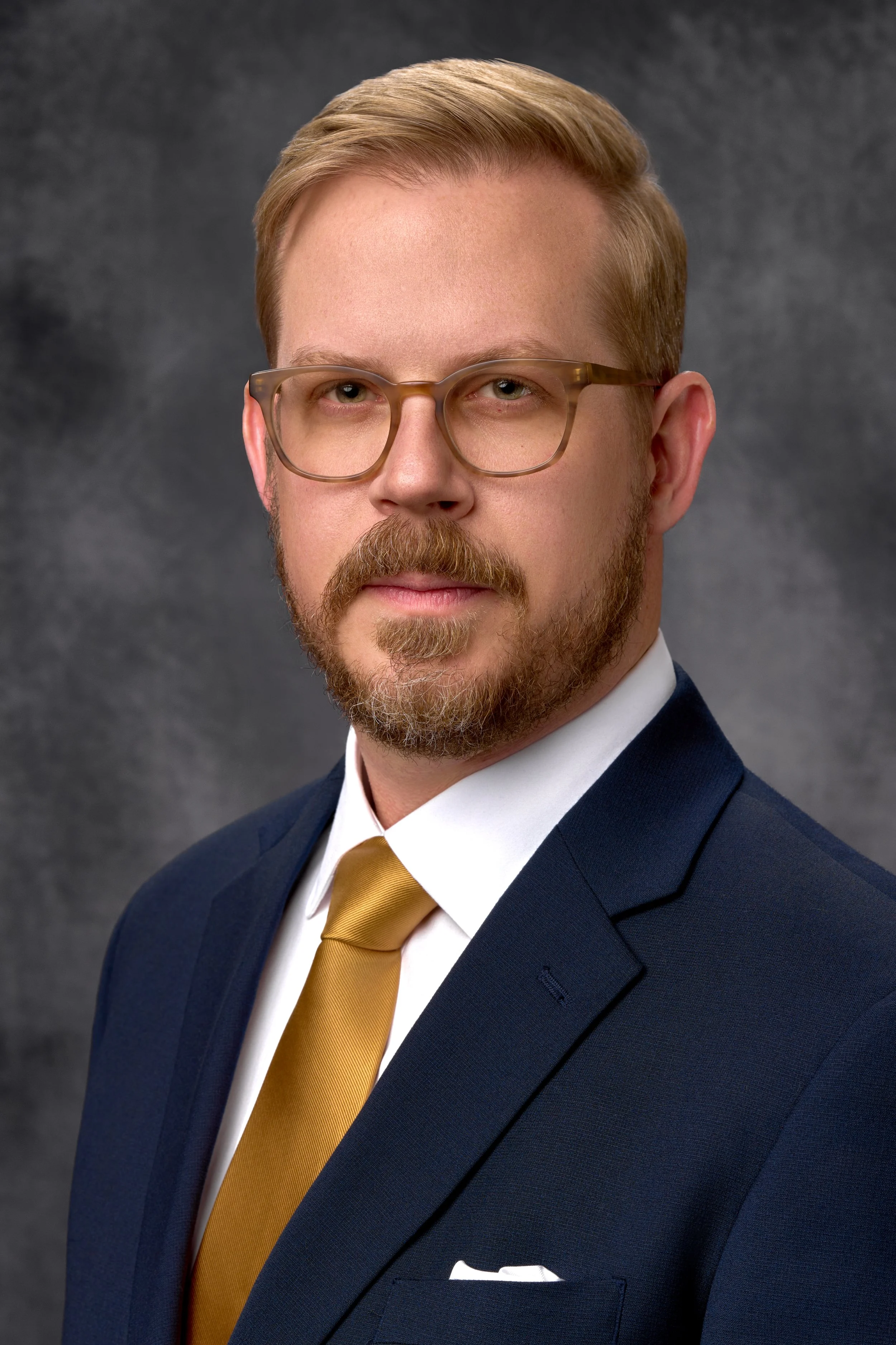 A professional headshot of a man with glasses, a beard, and light brown hair, wearing a navy blue suit, white shirt, and gold tie, against a dark gray background.