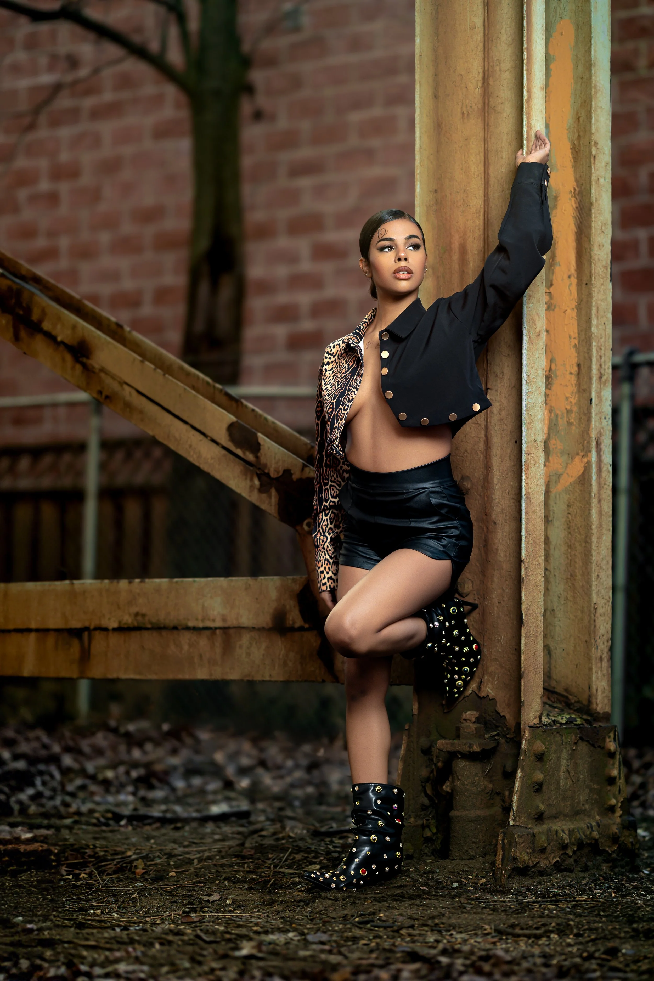 A woman with dark hair and makeup, wearing a draped leopard print and black jacket, black shorts, and studded black boots, posing outdoors against a rusted metal structure with a brick wall and trees in the background.