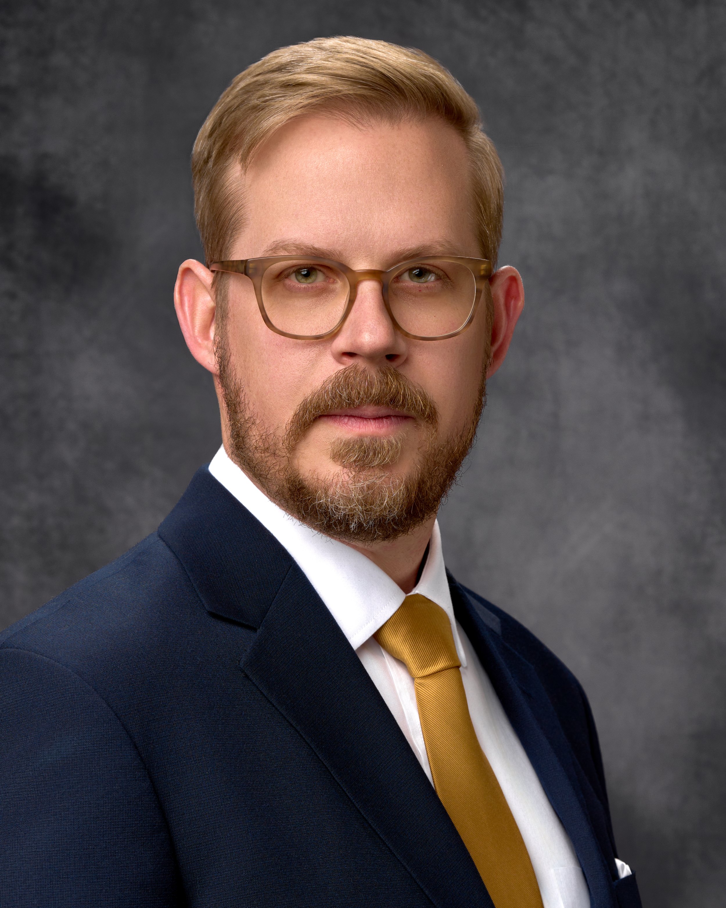 Chicago professional headshot portrait of business man with light brown hair, glasses, and a beard, dressed in a dark suit with a white shirt and yellow tie, posing against a gray textured background.