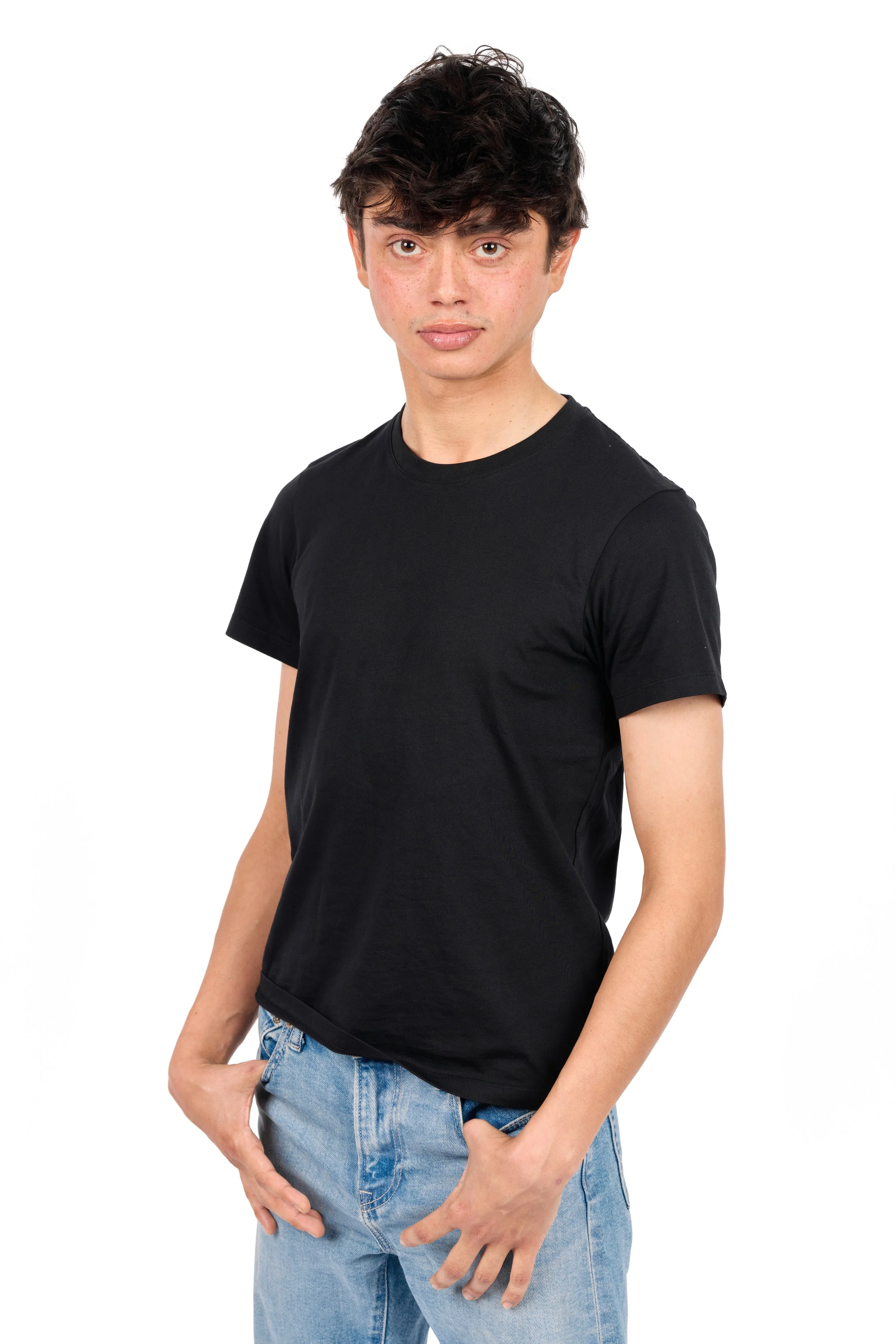 Young adult with dark wavy hair wearing a black t-shirt and light blue jeans, posing with one hand in pocket against a plain white background.