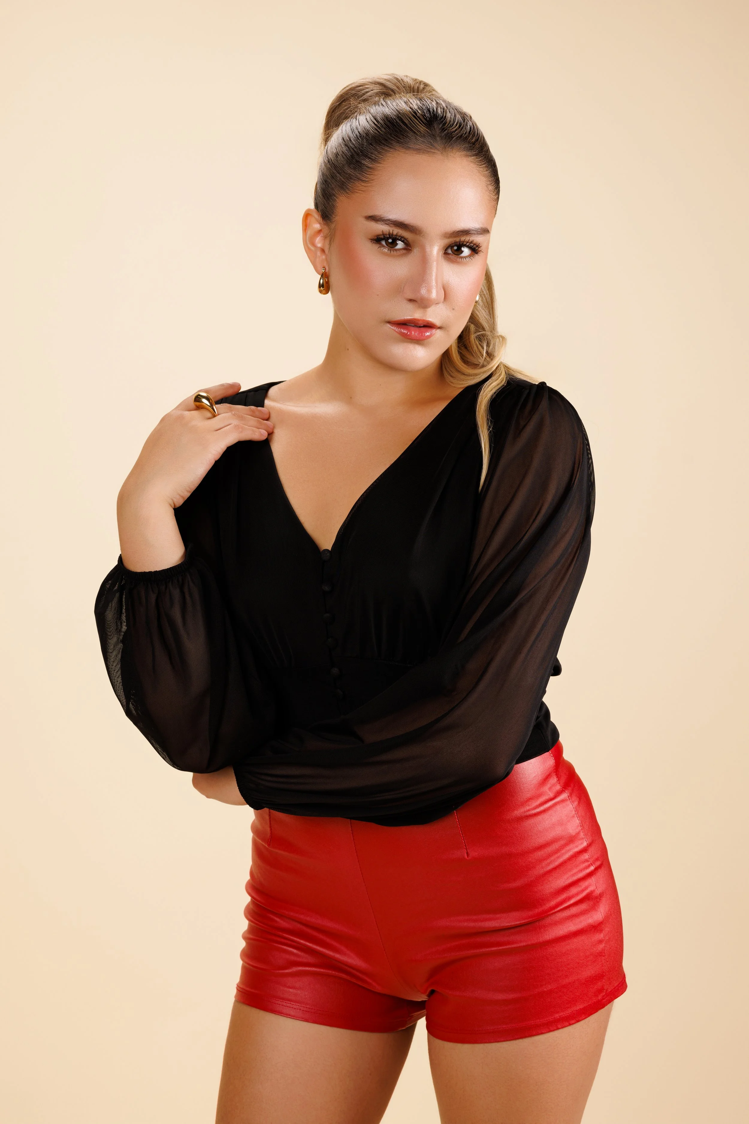 A woman with long blonde hair in a high ponytail, wearing gold hoop earrings, a black sheer long-sleeve blouse, and red leather shorts, standing against a beige background.