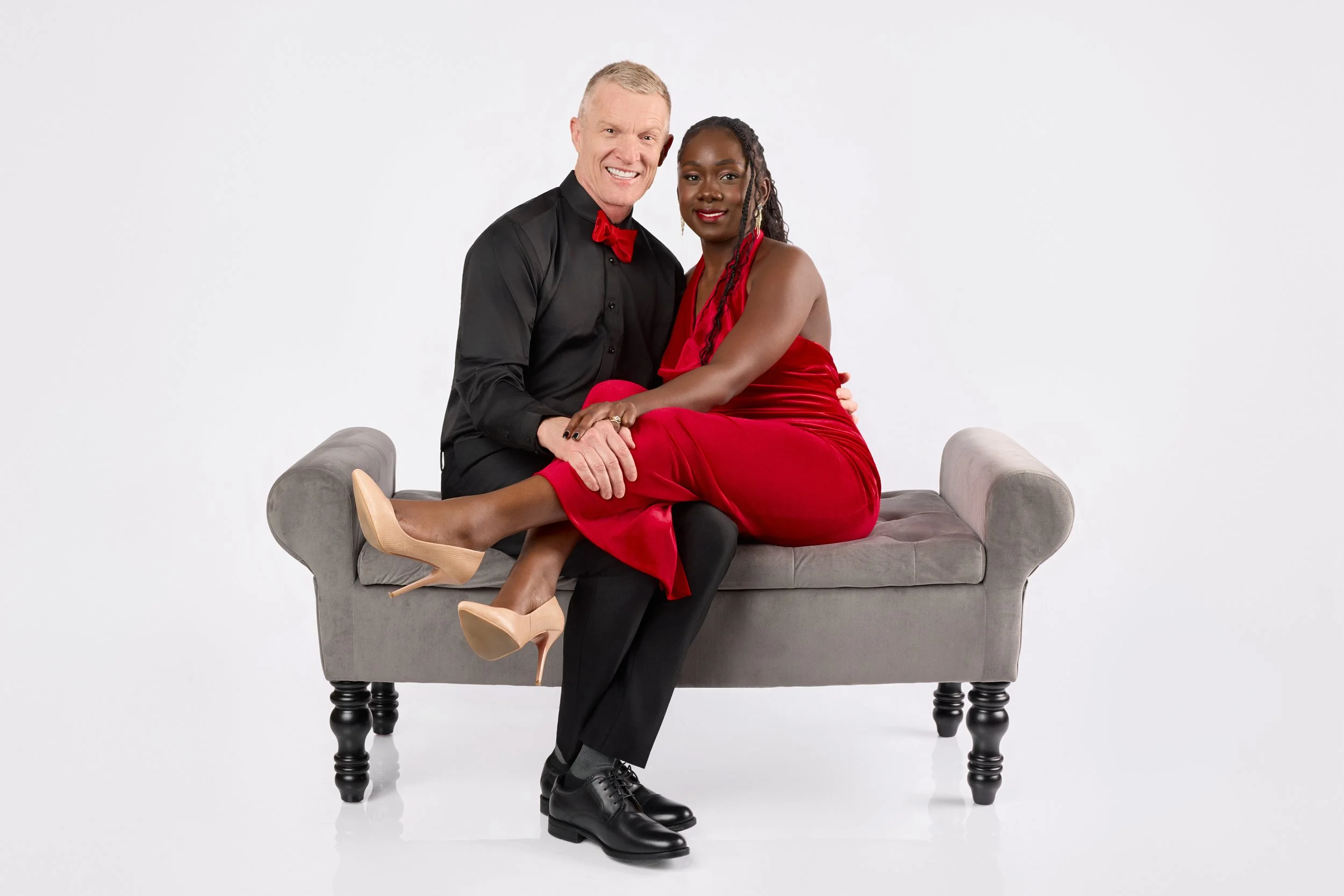 A smiling man in a black shirt with a red bow tie and a woman in a red dress sitting together on a grey loveseat against a white background.