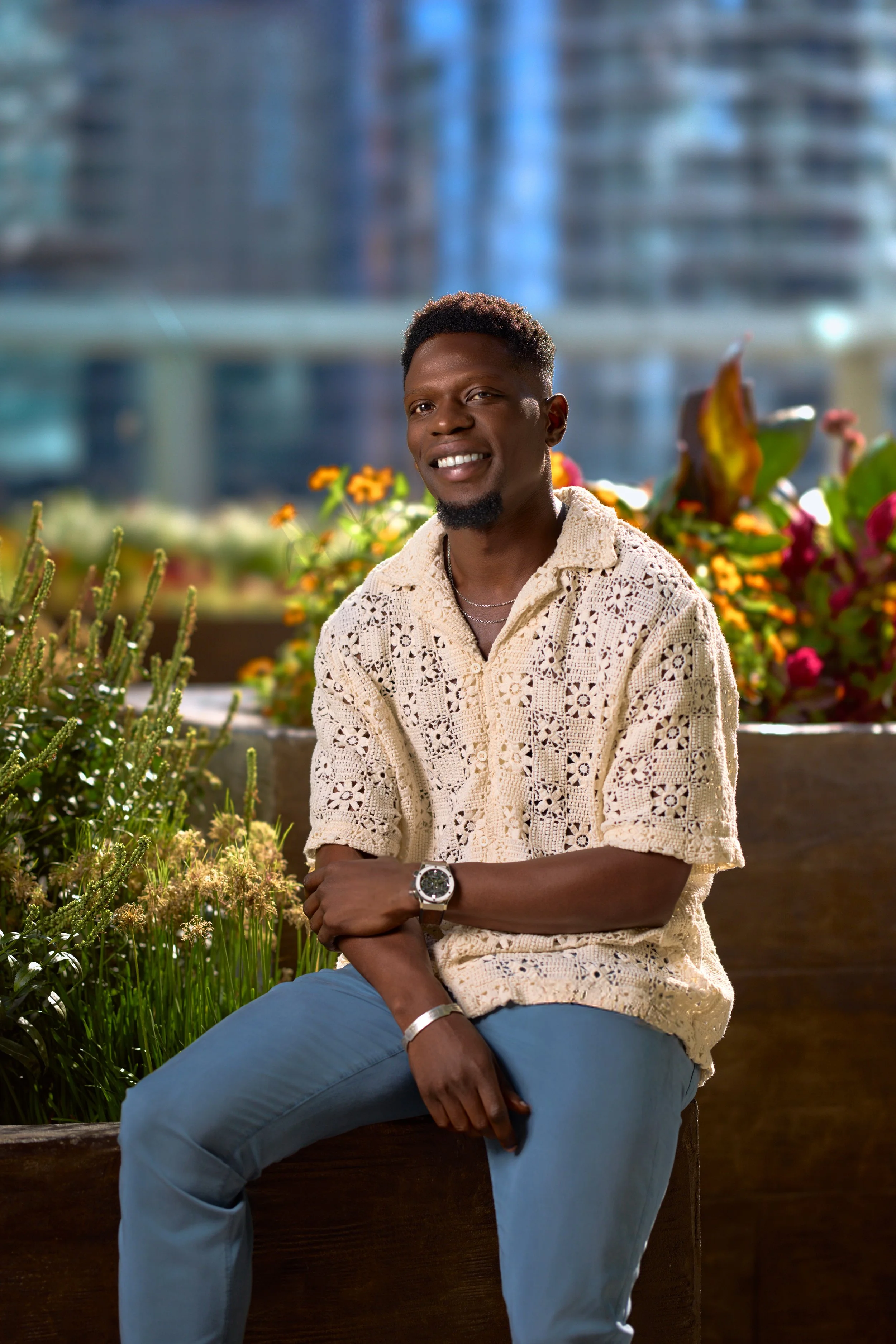 A man sitting outdoors near colorful flowers with a cityscape and glass building in the background, smiling and wearing a beige crocheted shirt, blue jeans, and accessories.