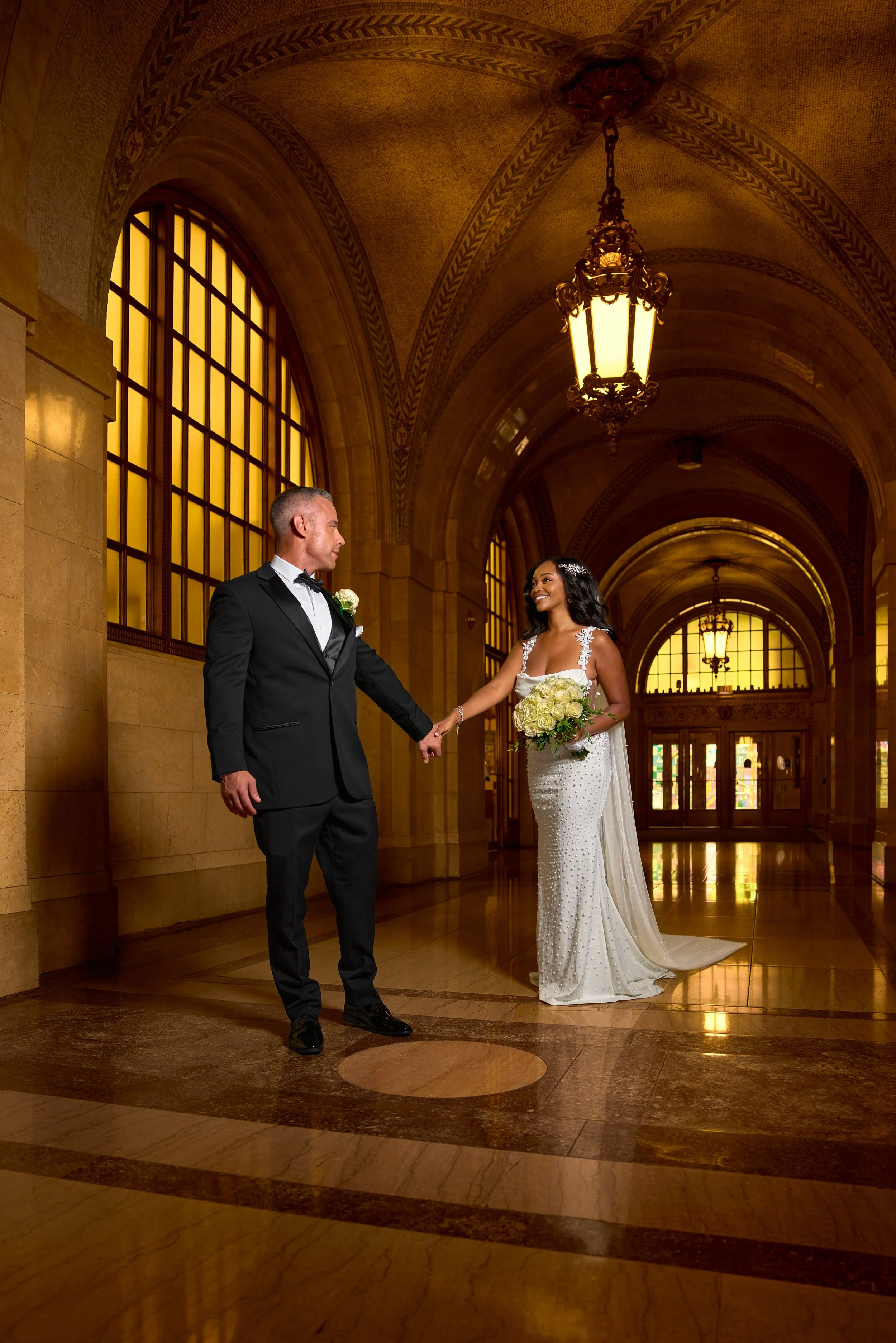 A bride and groom holding hands in an elegant, grand indoor setting with arched ceilings, large stained glass windows, and hanging chandeliers, celebrating their wedding.