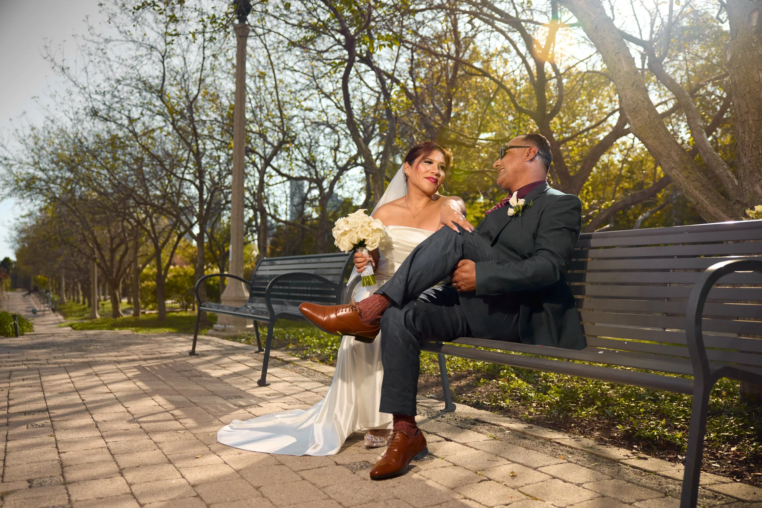 A bride and groom sitting on a park bench, smiling at each other during their wedding photos. The bride is wearing a white wedding dress and holding a bouquet, while the groom is in a black suit with brown shoes. They are surrounded by trees with sunlight filtering through the branches.