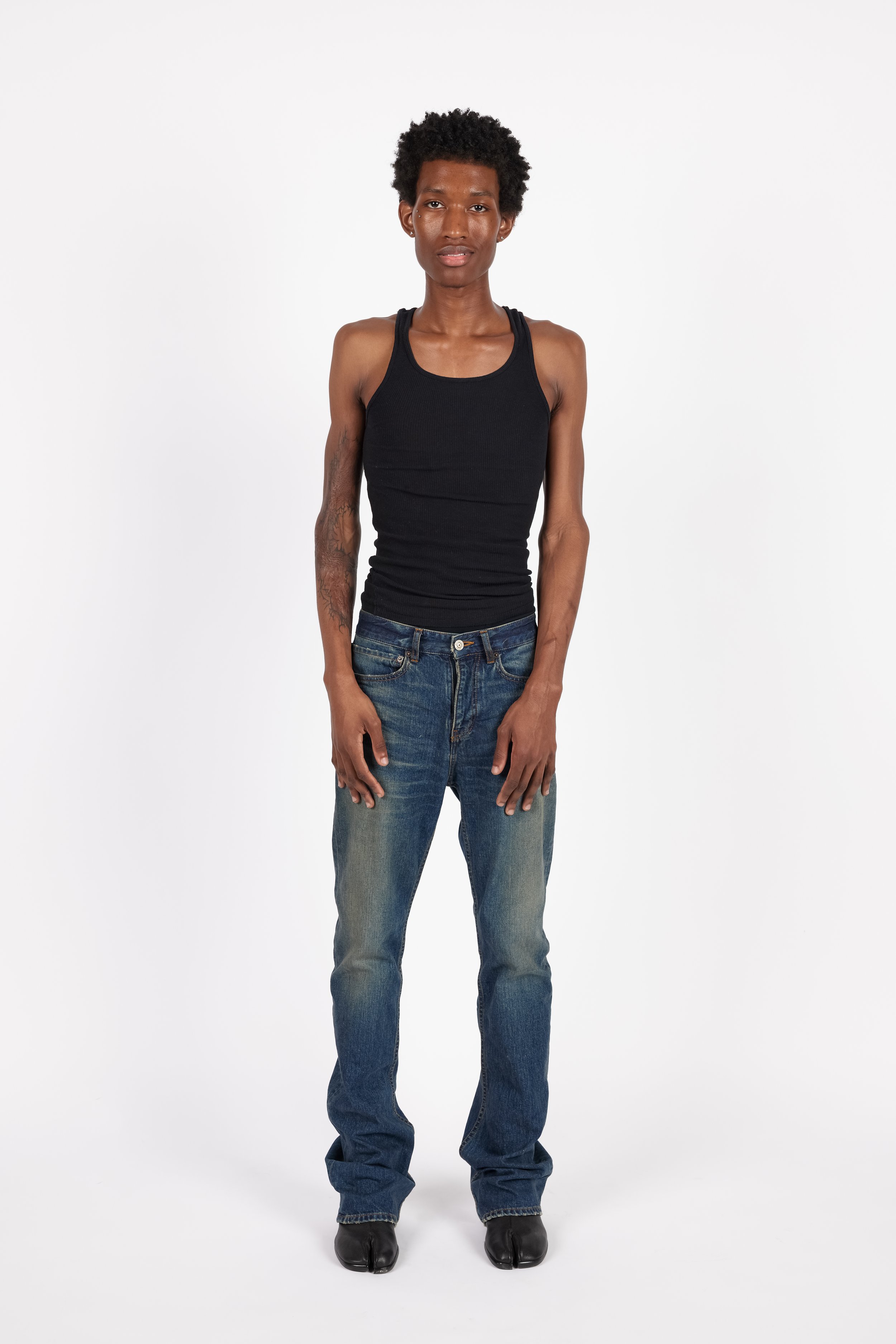 Chicago Modelling Digital of a young man with short curly hair standing against a plain white background, wearing a black tank top, blue jeans, and black shoes.
