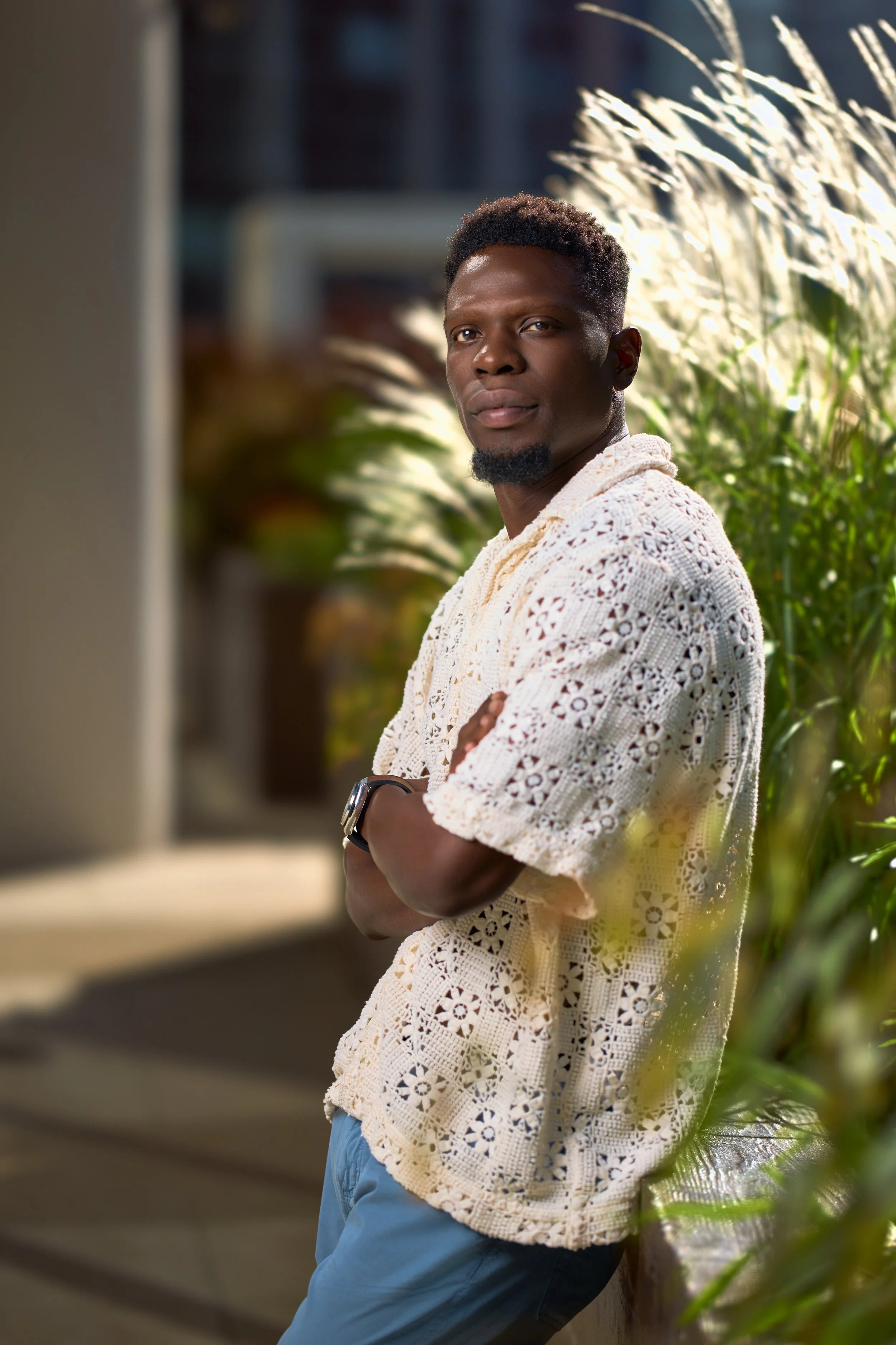 A young man with a dark complexion, short curly hair, and a beard, standing outdoors next to green plants. He is wearing a white crocheted shirt, blue pants, and a wristwatch, with arms crossed and looking at the camera.