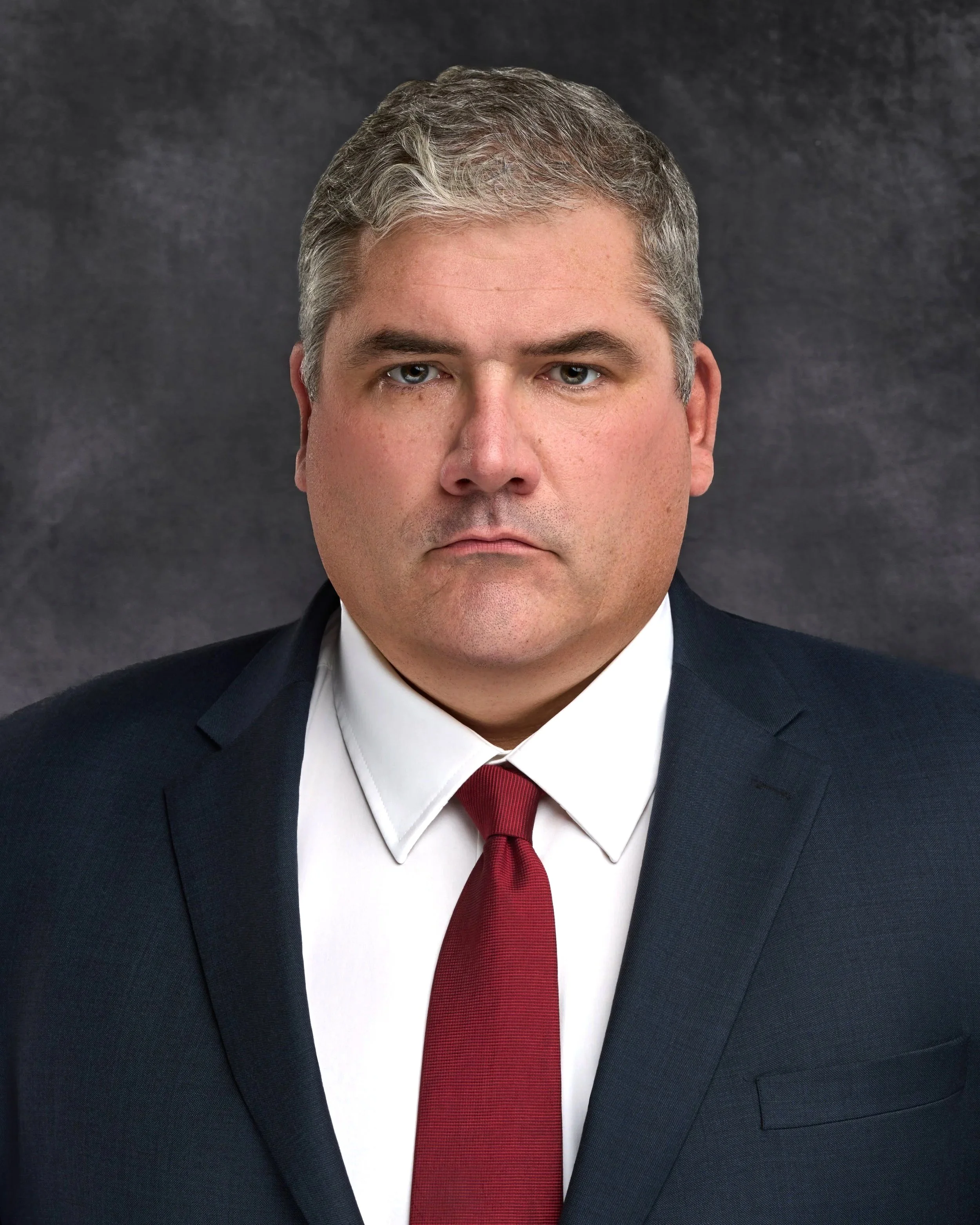 Chicago professional headshot portrait of a corporate man, a middle-aged man in a suit, white shirt, and red tie, with short gray hair, against a dark textured background.