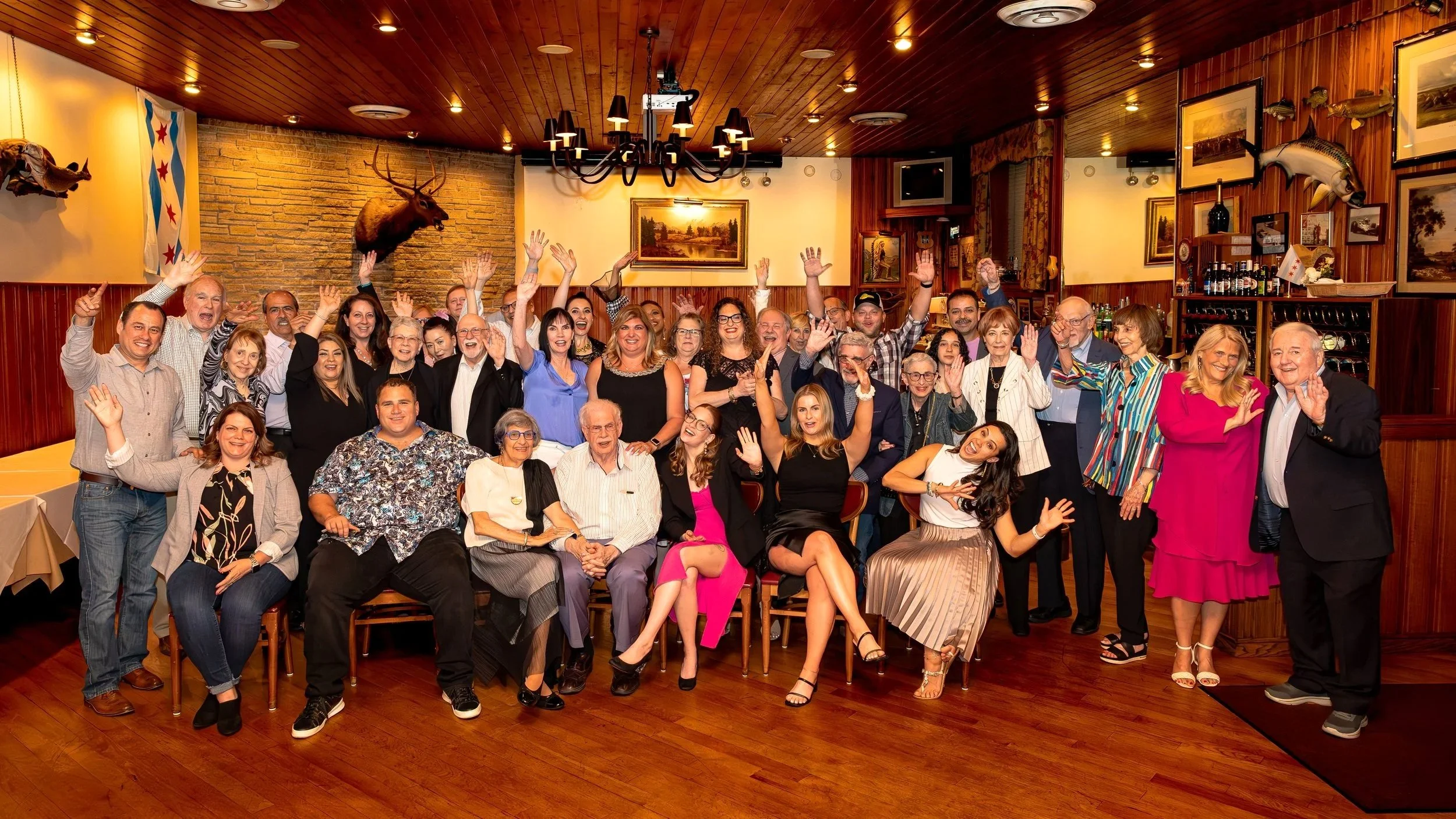 Group of people gathered in a restaurant, smiling and waving at the camera, with wooden walls decorated with paintings and mounted animals.