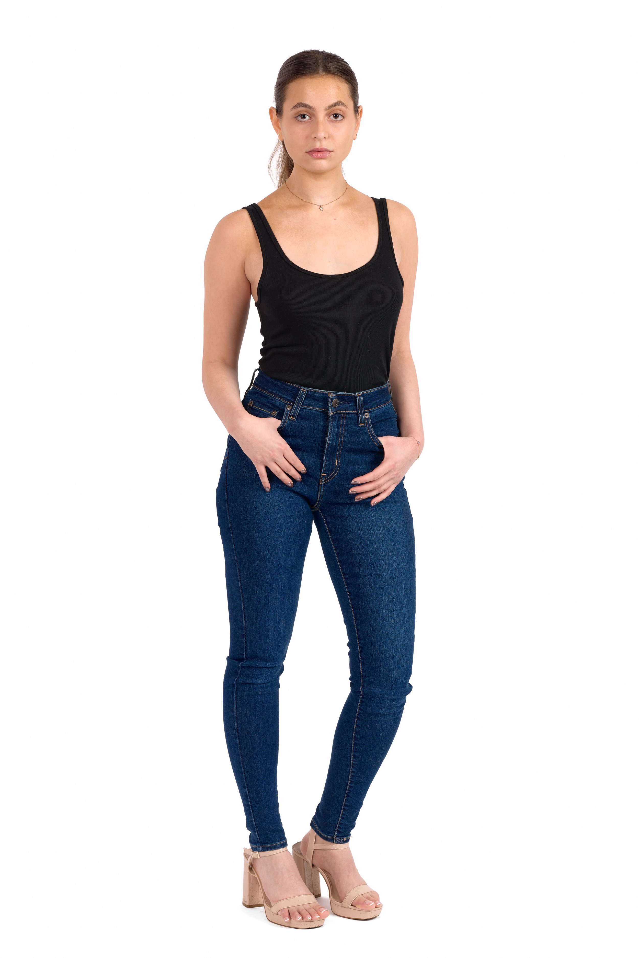 Chicago Modelling Digital of a young woman with brown hair tied back, wearing a black tank top, high-waisted blue jeans, and beige open-toe heels, standing with one hand in her pocket against a white background.