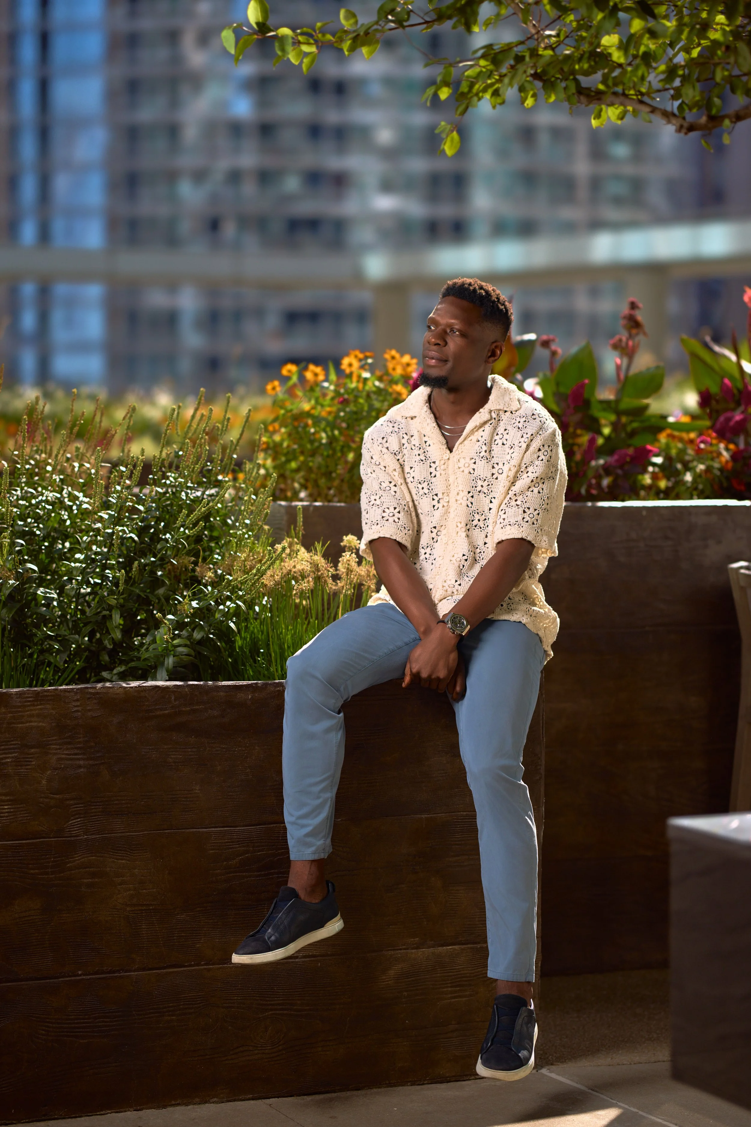 Chicago fashion photoshoot of a male model sitting on a wooden ledge outside, surrounded by flowering plants and trees, with a cityscape in the background.