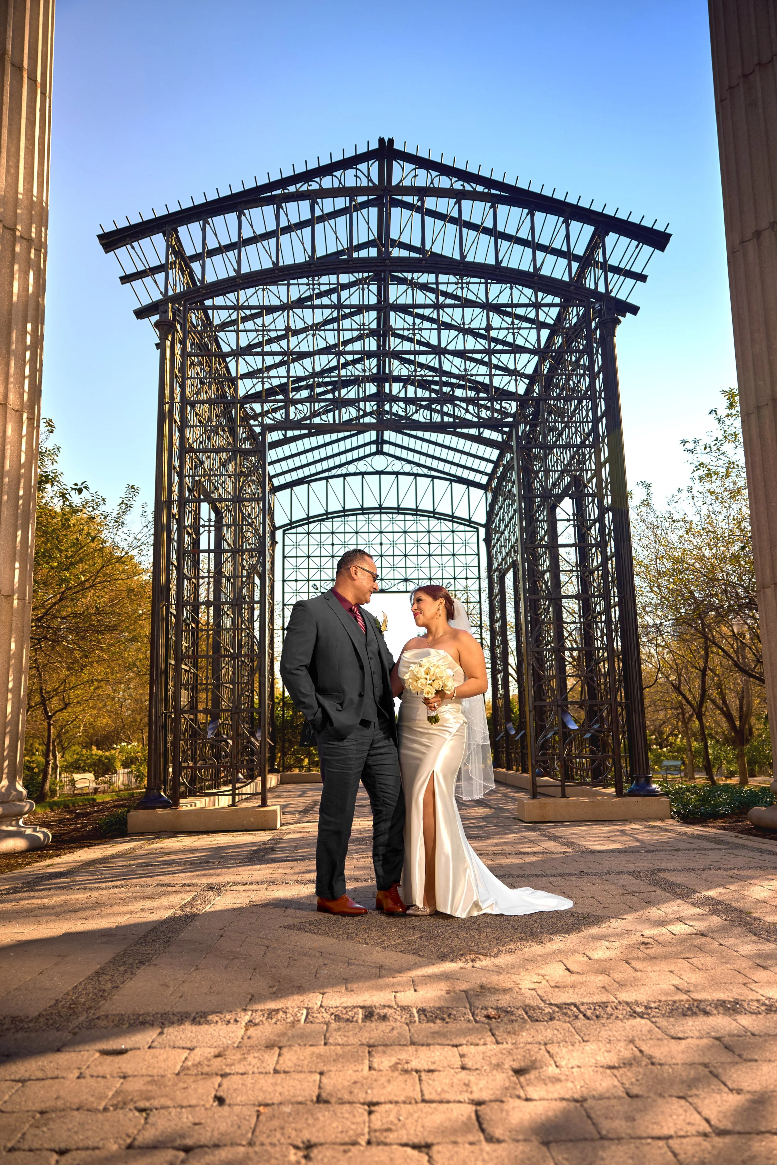 Engagement portraits with the bride and groom standing under a decorative metal archway, looking at each other, with trees in the background and sunlight casting shadows on the brick ground.