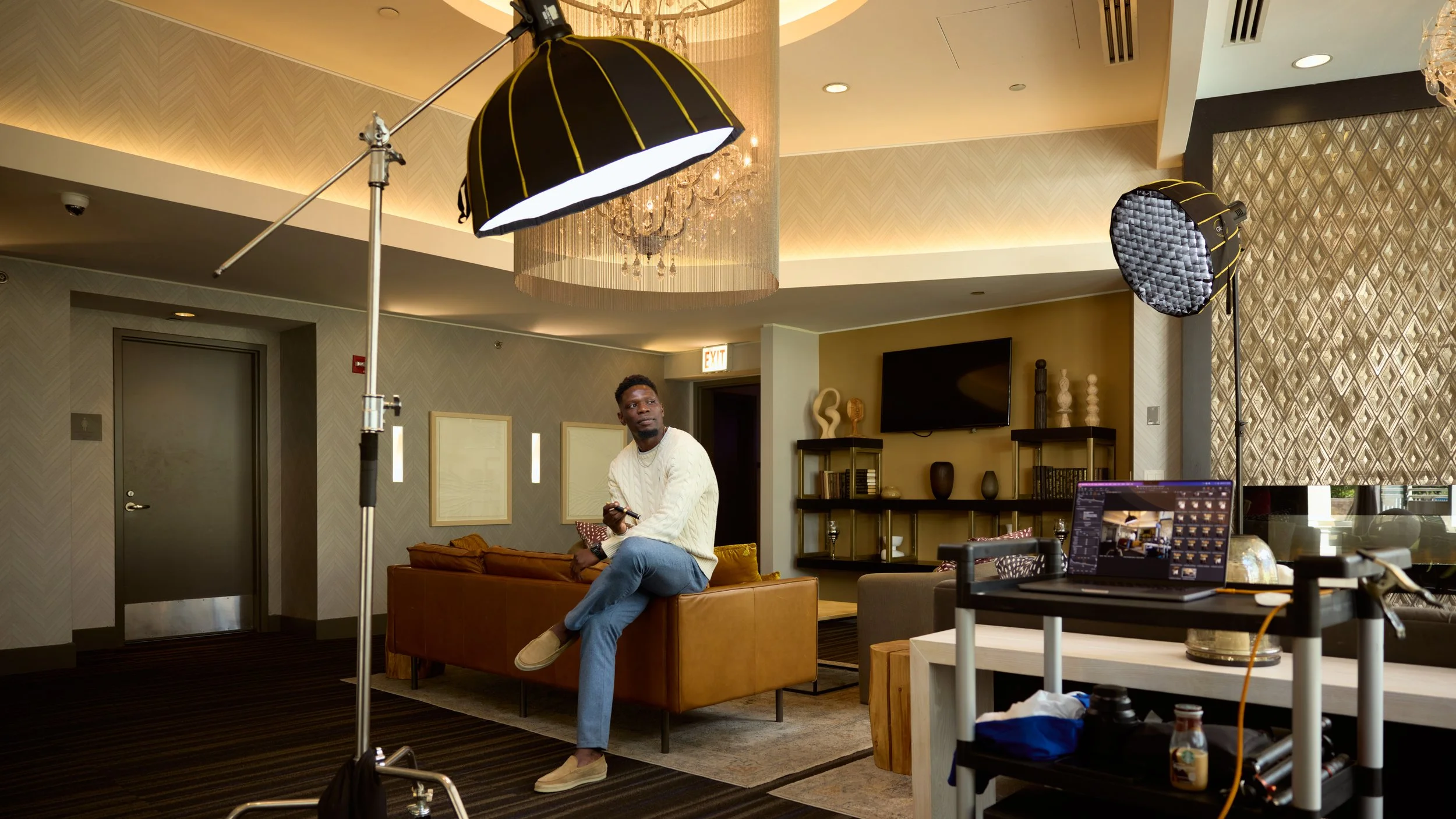 A man sits on the back of a brown leather couch in a modern, well-decorated living room with a large chandelier and two studio lights on stands. There is a large flat-screen TV on the wall, and a shelf with decorative objects behind him. A laptop on a cart and some equipment are in the foreground.