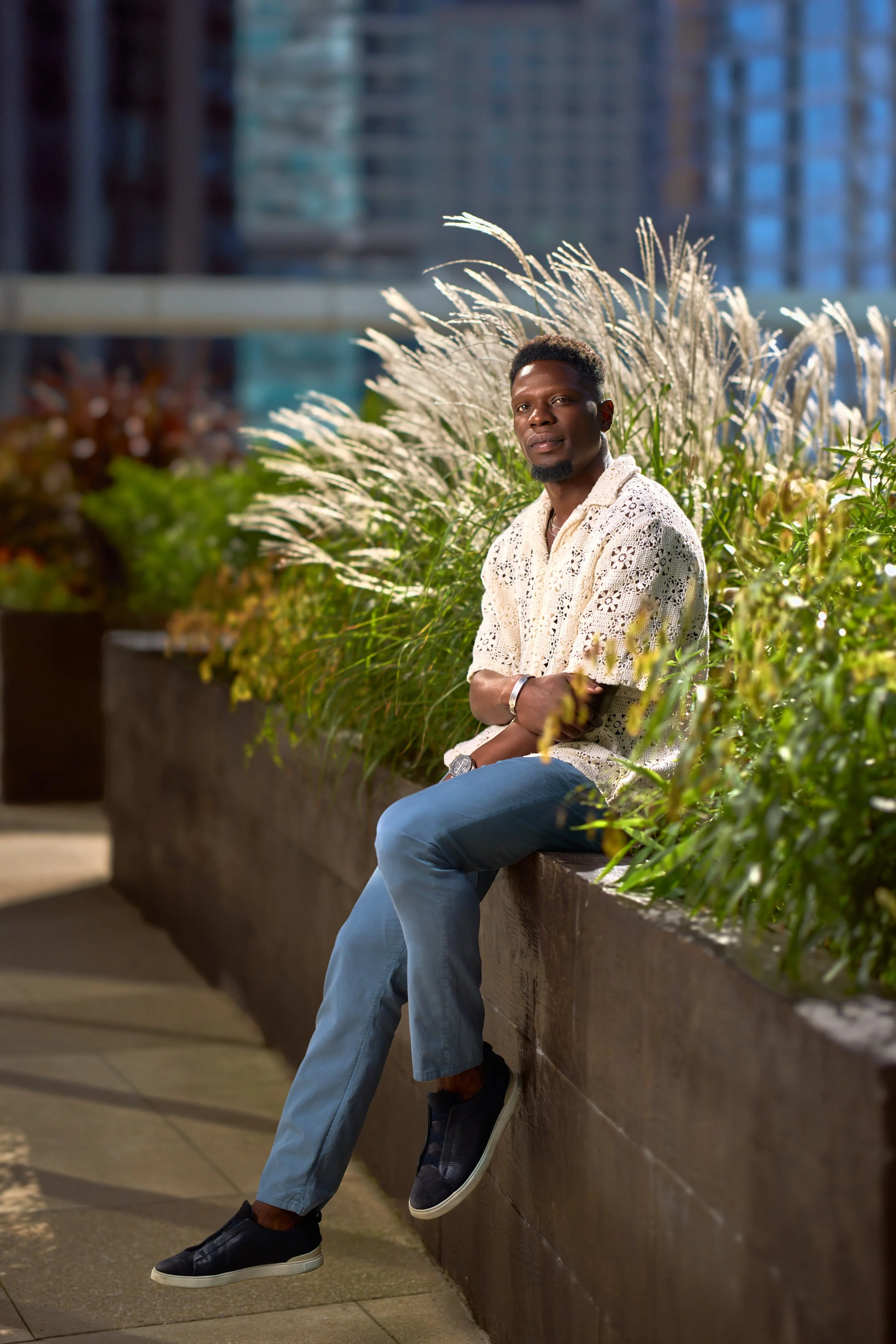 Chicago fashion photoshoot of a male model sitting on a ledge in an outdoor urban setting with tall grasses and greenery, wearing a cream-colored crochet shirt, light blue pants, and black sneakers, during evening or night.