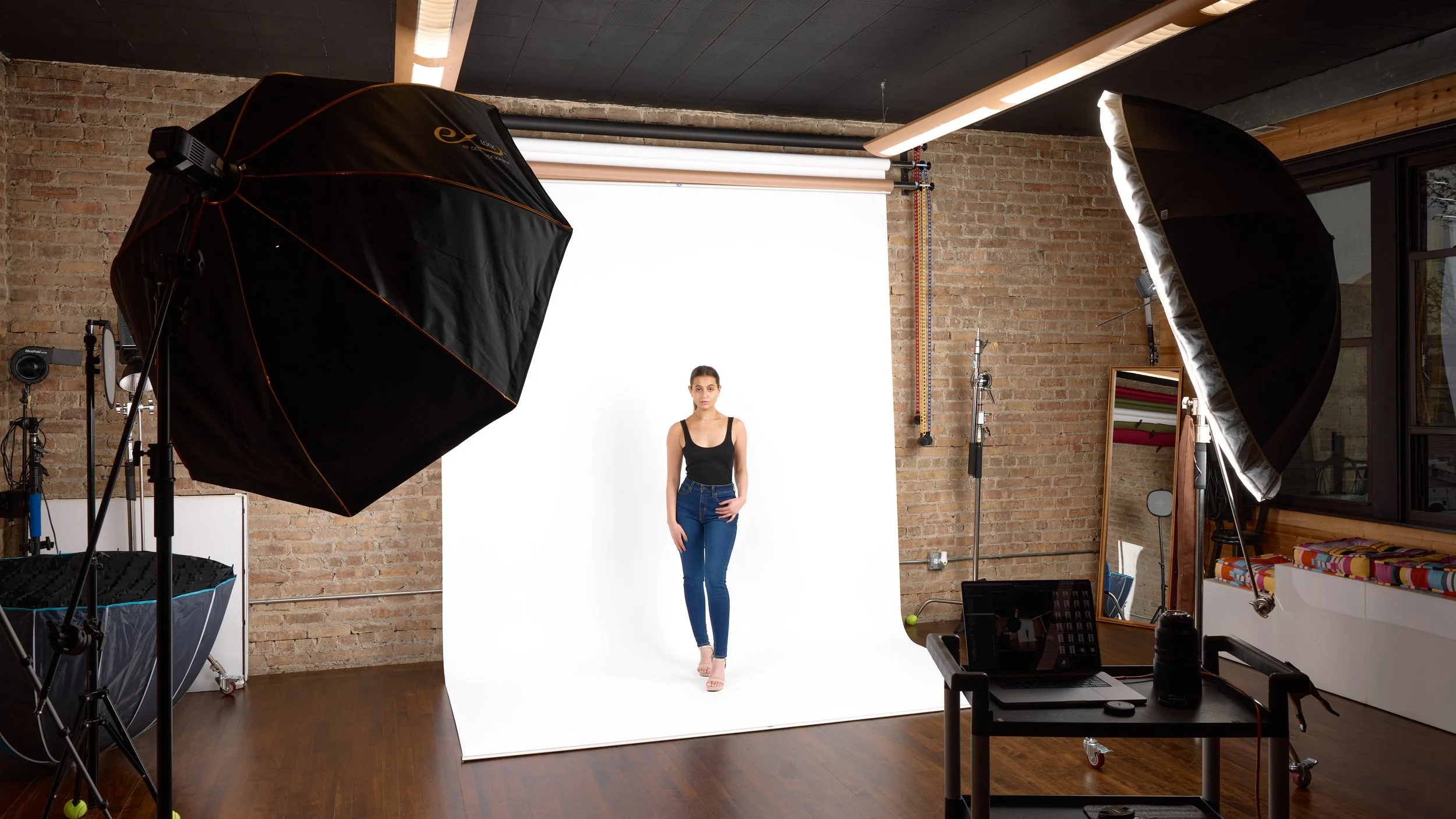Model standing in photography studio with white backdrop, photography equipment, and softbox lights.