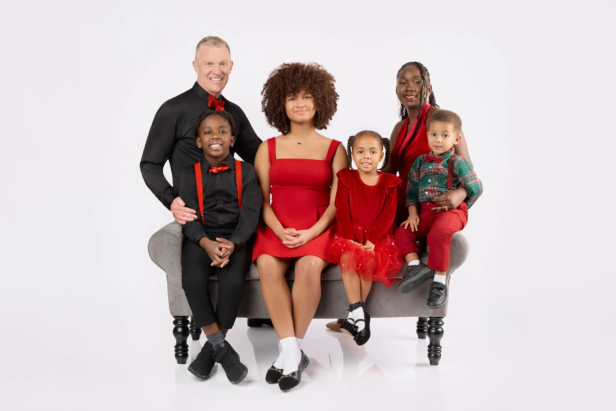 Family of six dressed in black and red, sitting on a gray couch against a white background.
