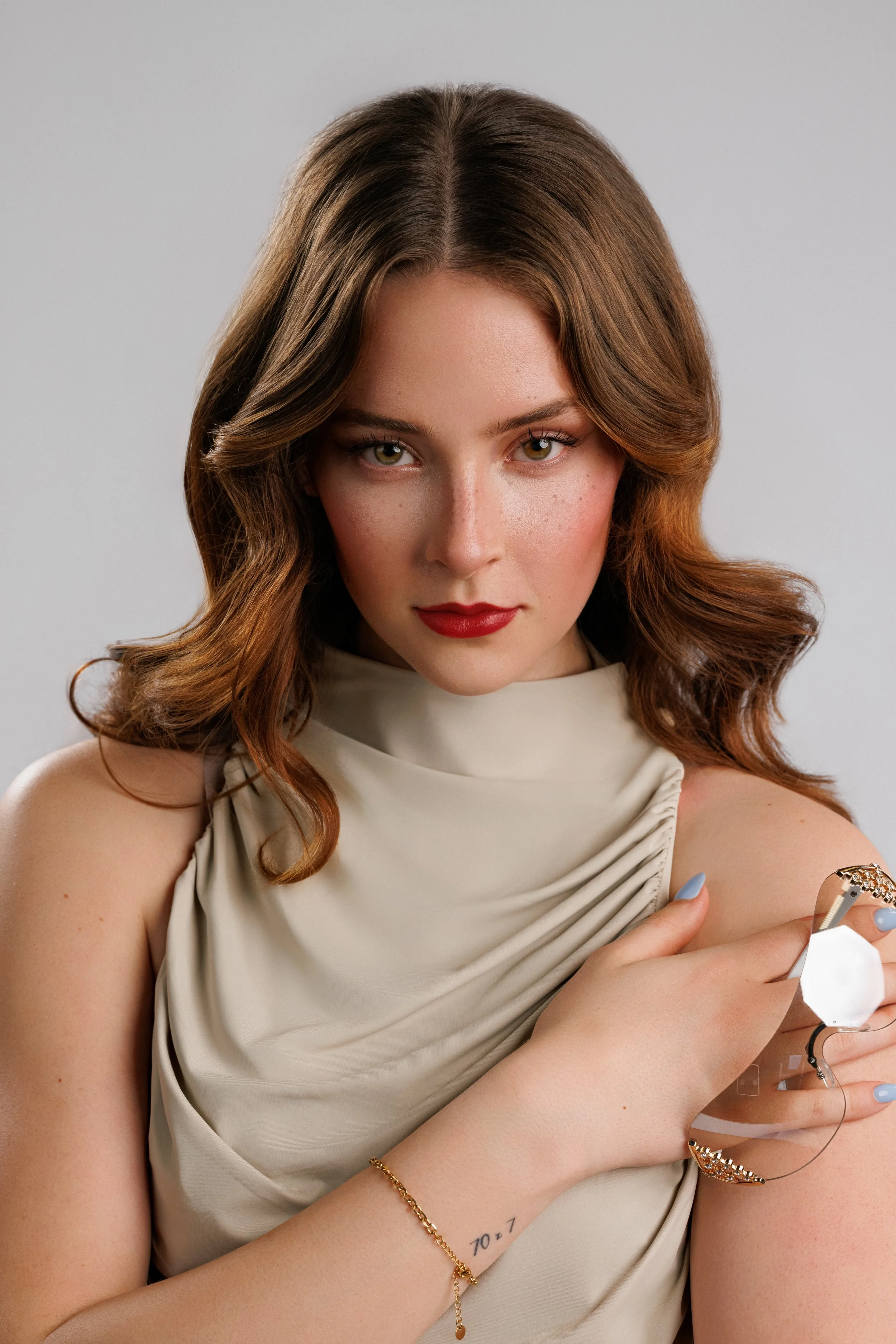 A woman with wavy brown hair, wearing a cream-colored sleeveless top, holding a cigarette with a silver ring and gold bracelet, looking directly at the camera