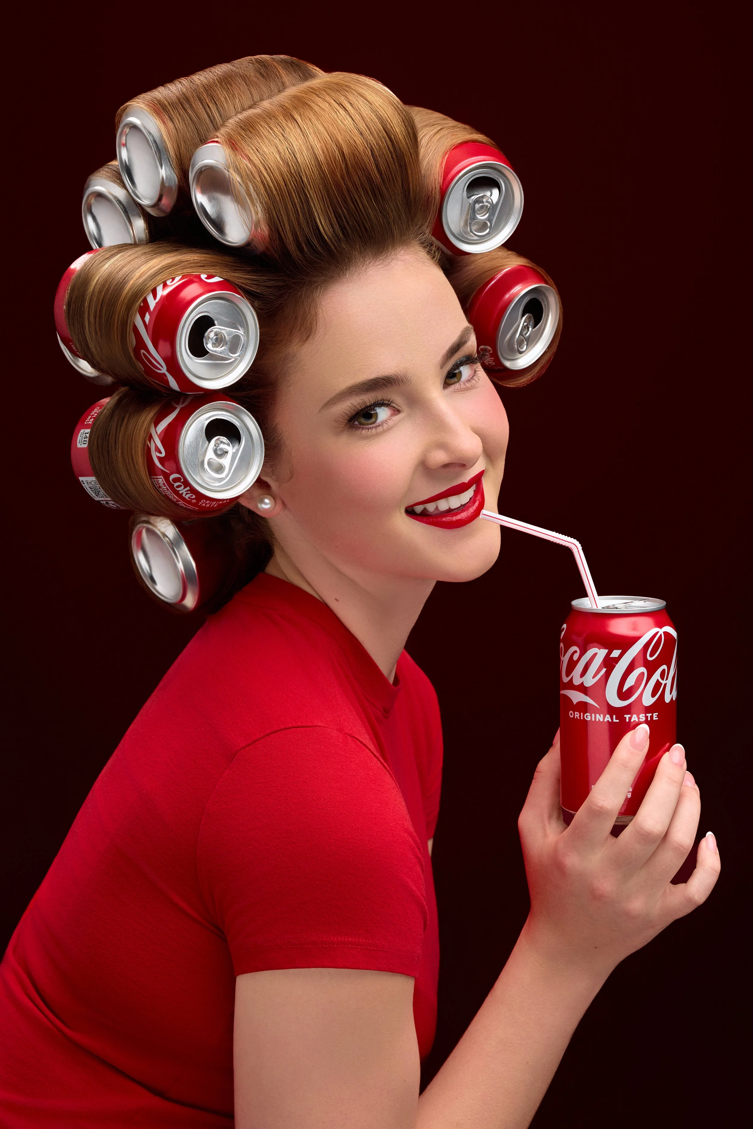 Creative branding photoshoot of a woman with hair styled in large curls made from Coca-Cola cans, drinking from a Coca-Cola can using a straw, against a dark red background.