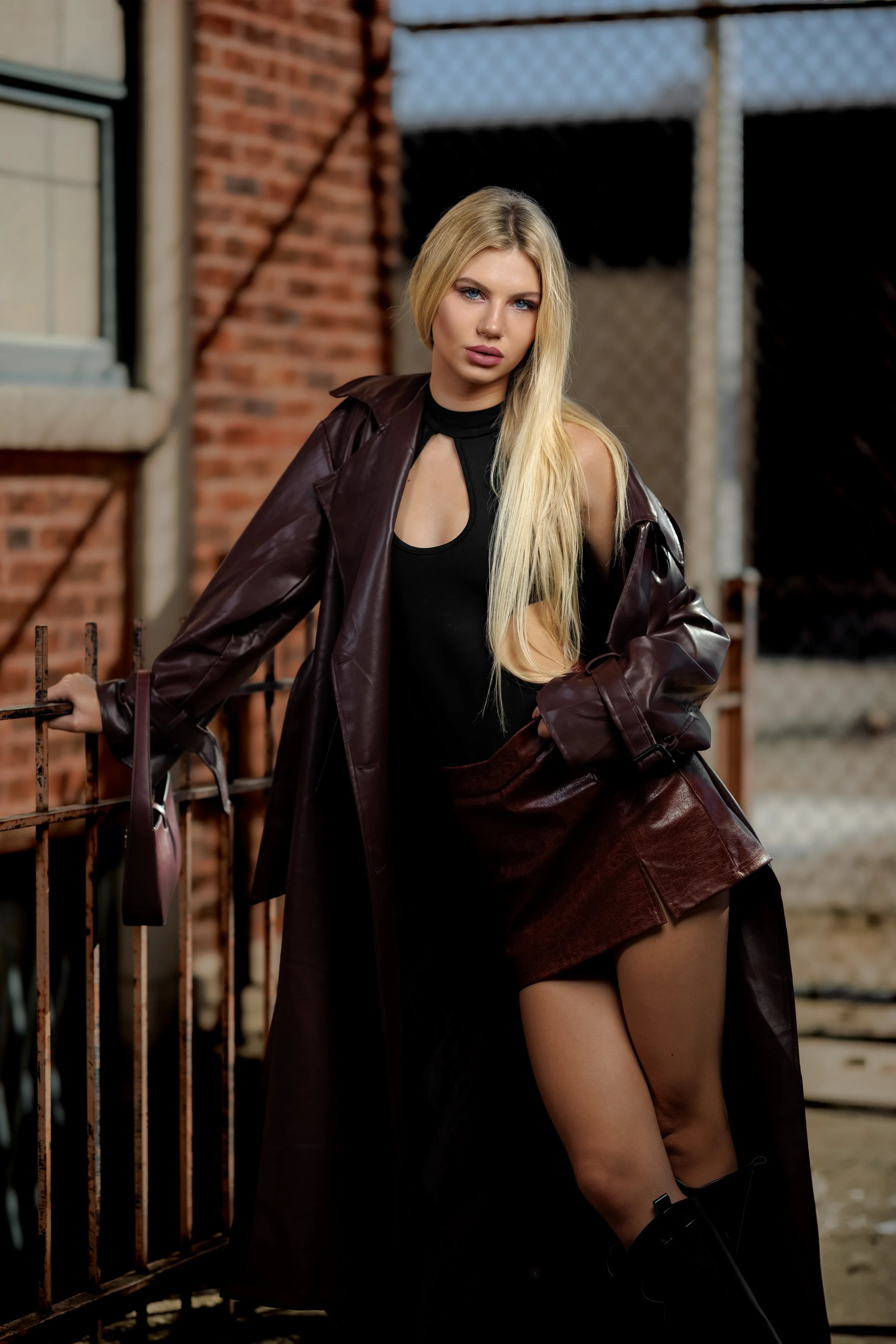 A woman with long blonde hair wearing a black top, a brown leather skirt, black boots, and a long brown leather coat, leaning against a metal fence outdoors with a brick building in the background.