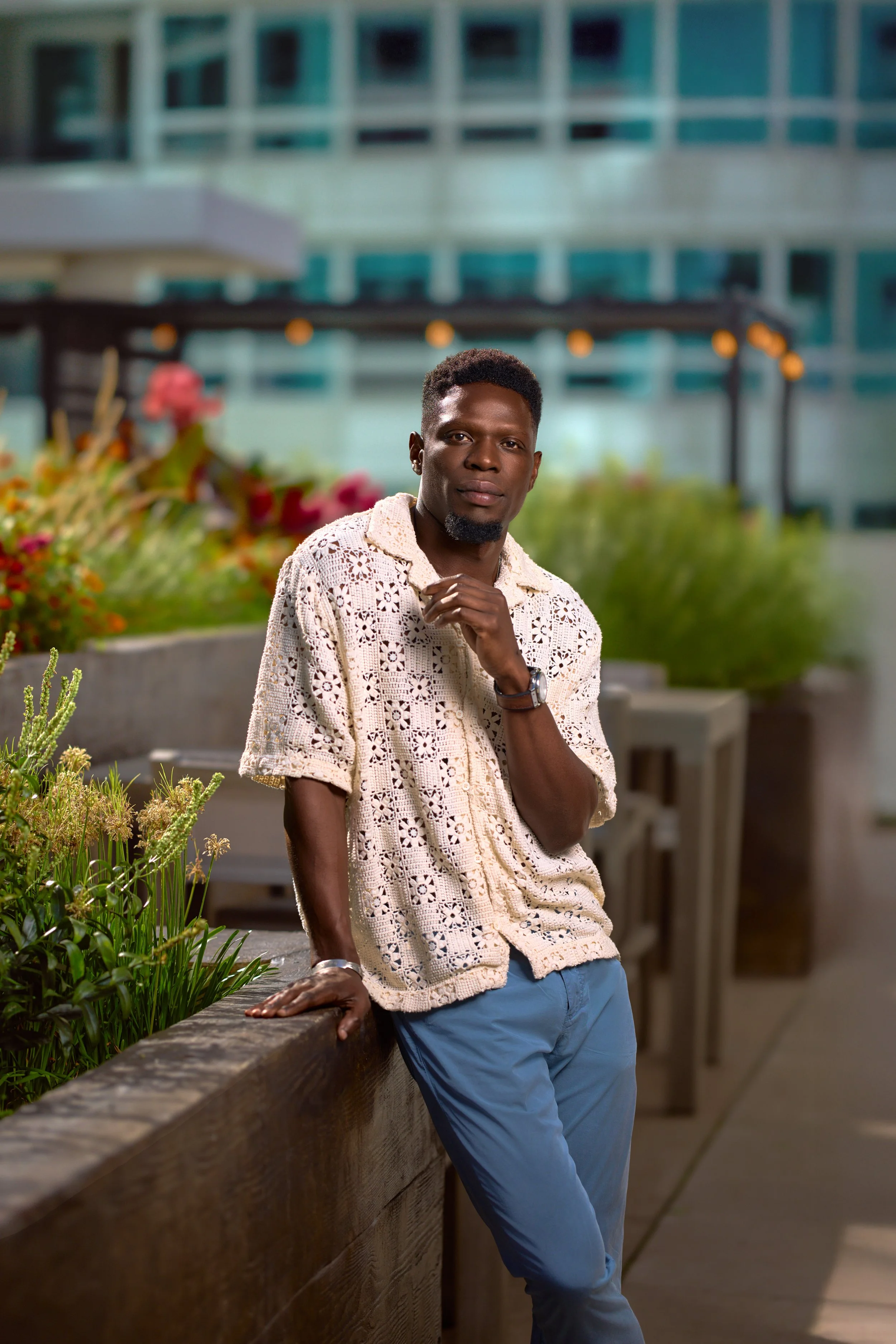 A man wearing a cream crochet shirt and blue pants poses outdoors at night, leaning on a wooden railing surrounded by plants and flowers with a modern building in the background.