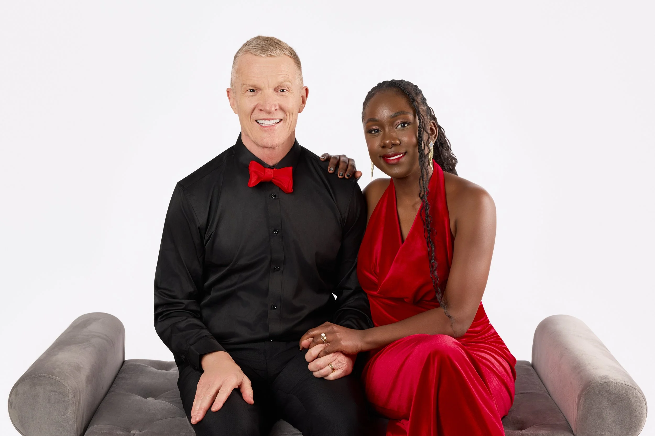 A man and woman sitting close on a grey bench against a white background. The man is wearing a black shirt and red bow tie, smiling. The woman is wearing a red dress, smiling, with her hand on the man's knee and her arm around his shoulder.