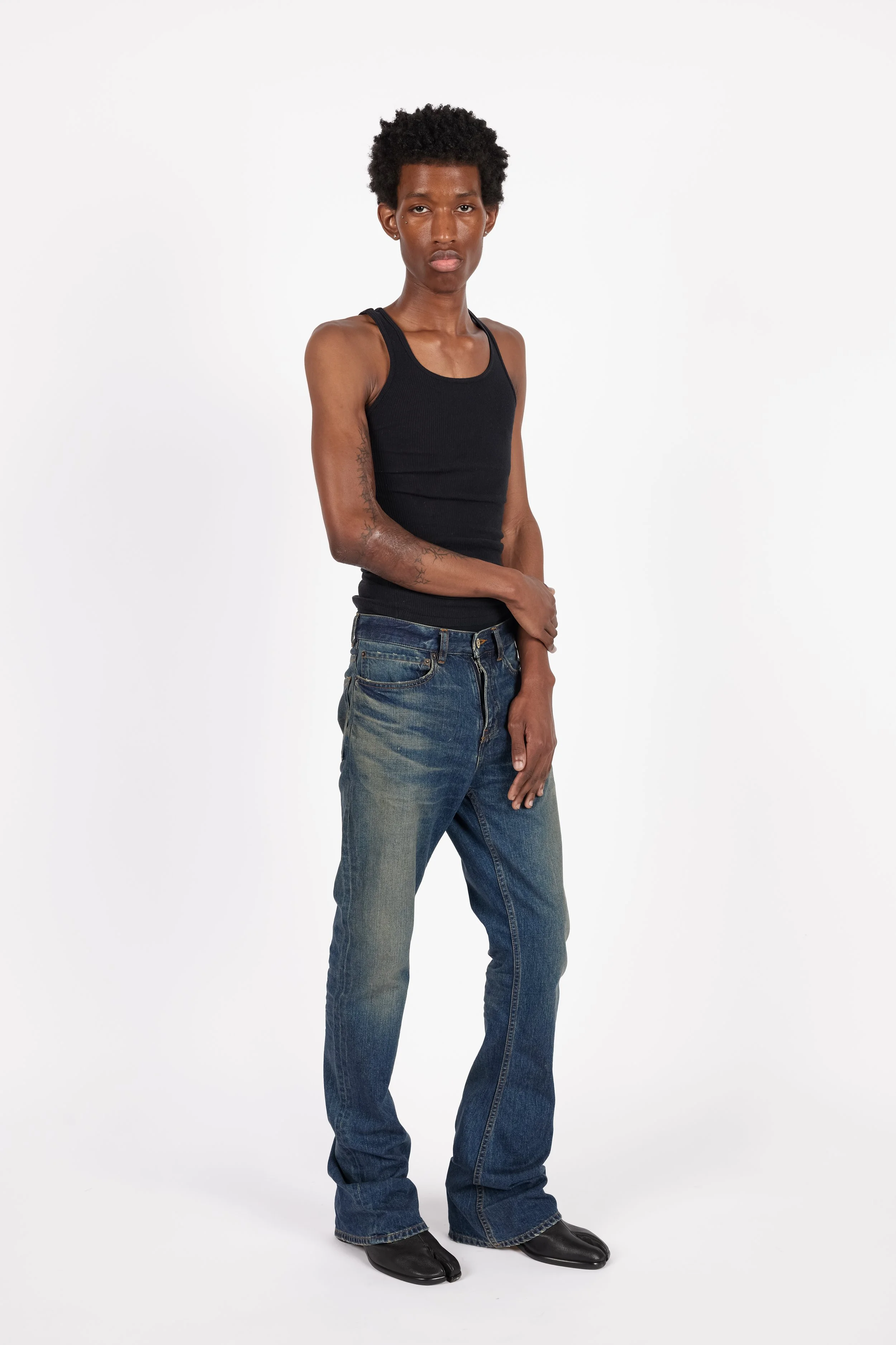 Chicago Modelling Digital of a young man with short curly hair wearing a black tank top, blue jeans, and black shoes standing against a plain white background.