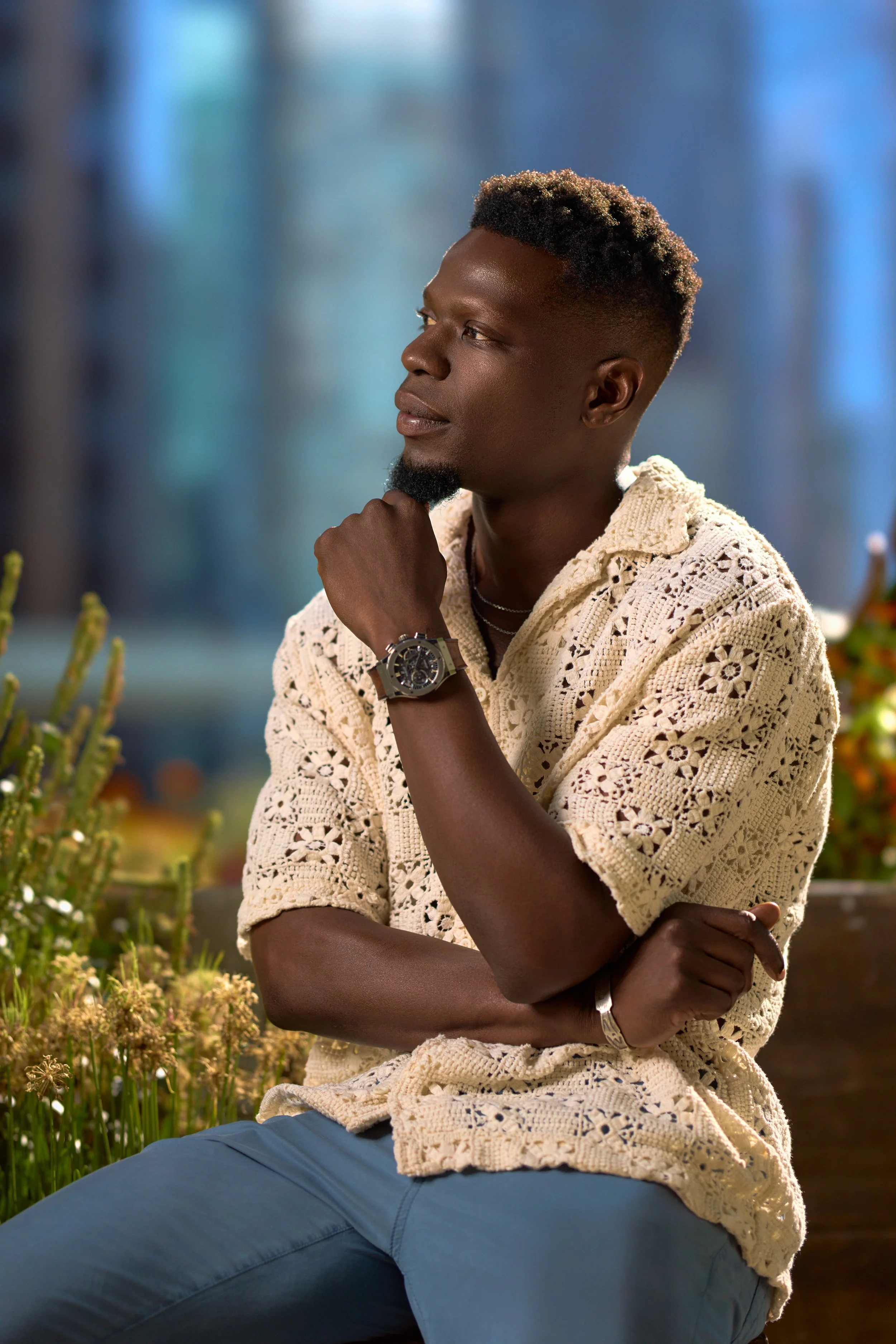 A young man with dark skin, short curly hair, and a goatee is sitting outdoors, wearing a cream-colored crocheted sweater over a black shirt, light blue pants, and a wristwatch. He is resting his chin on his right hand with a thoughtful expression.