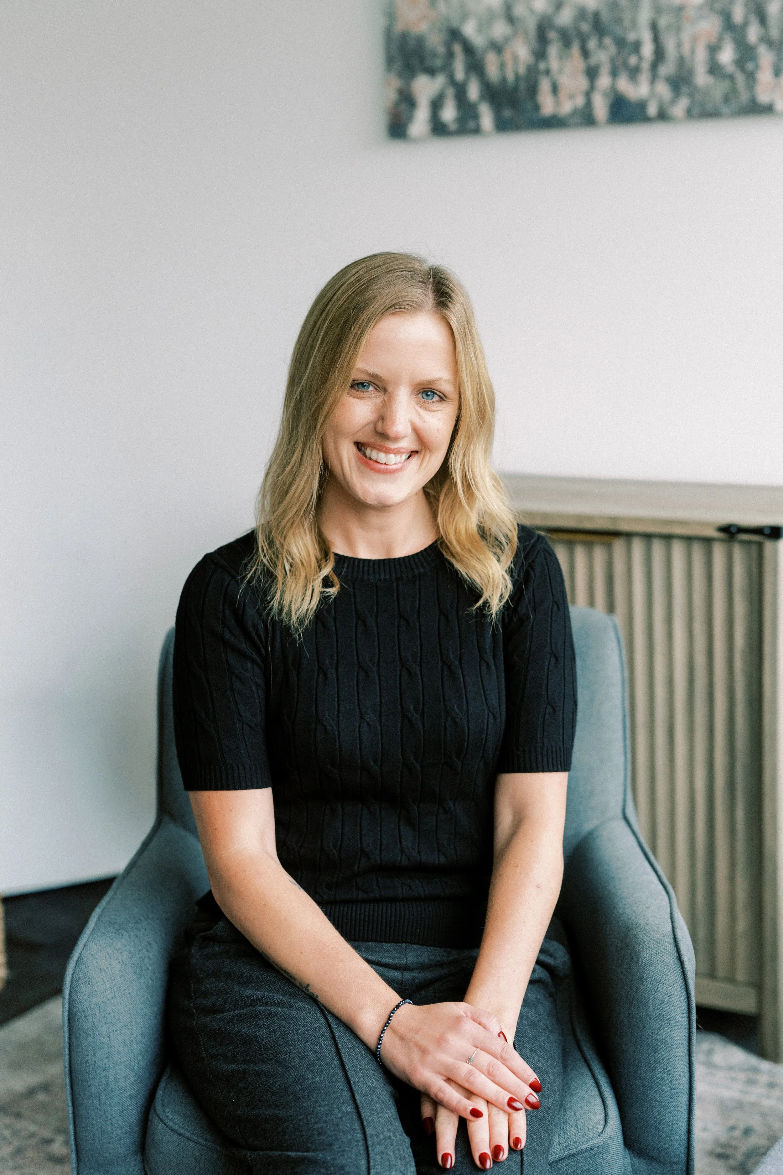 A woman with blond hair and blue eyes is sitting in a gray armchair, smiling at the camera. She is wearing a black, short-sleeved, cable-knit sweater and gray pants. Her hands are resting on her lap, with red painted nails and a bracelet on her left wrist. The background features a white wall with a colorful abstract painting and a wooden radiator cover.