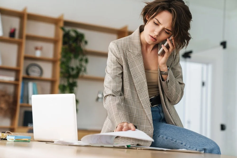 A woman with short, wavy brown hair, wearing a beige blazer and jeans, is sitting on a desk in an office while talking on a phone and looking at documents.
