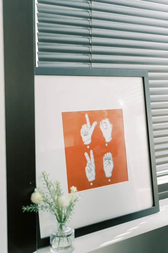 Framed print with four hand gesture illustrations on an orange background, placed on a windowsill beside a small vase with white flowers.