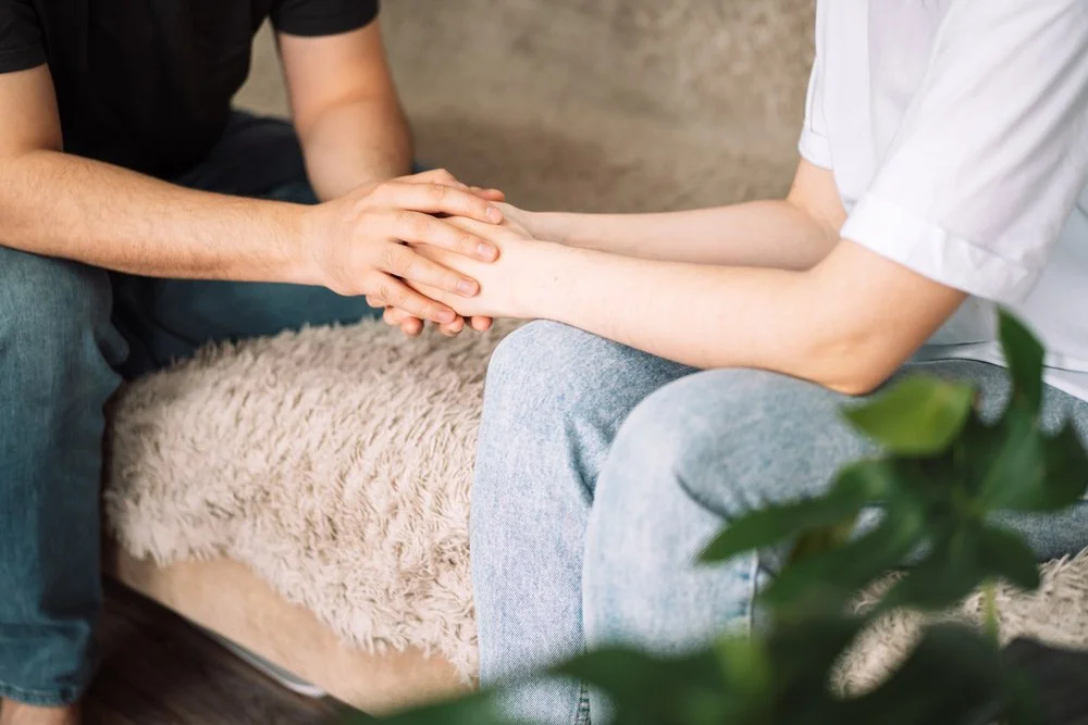 Two people sitting on a beige furry pouf, holding hands in a comforting manner in an indoor setting.