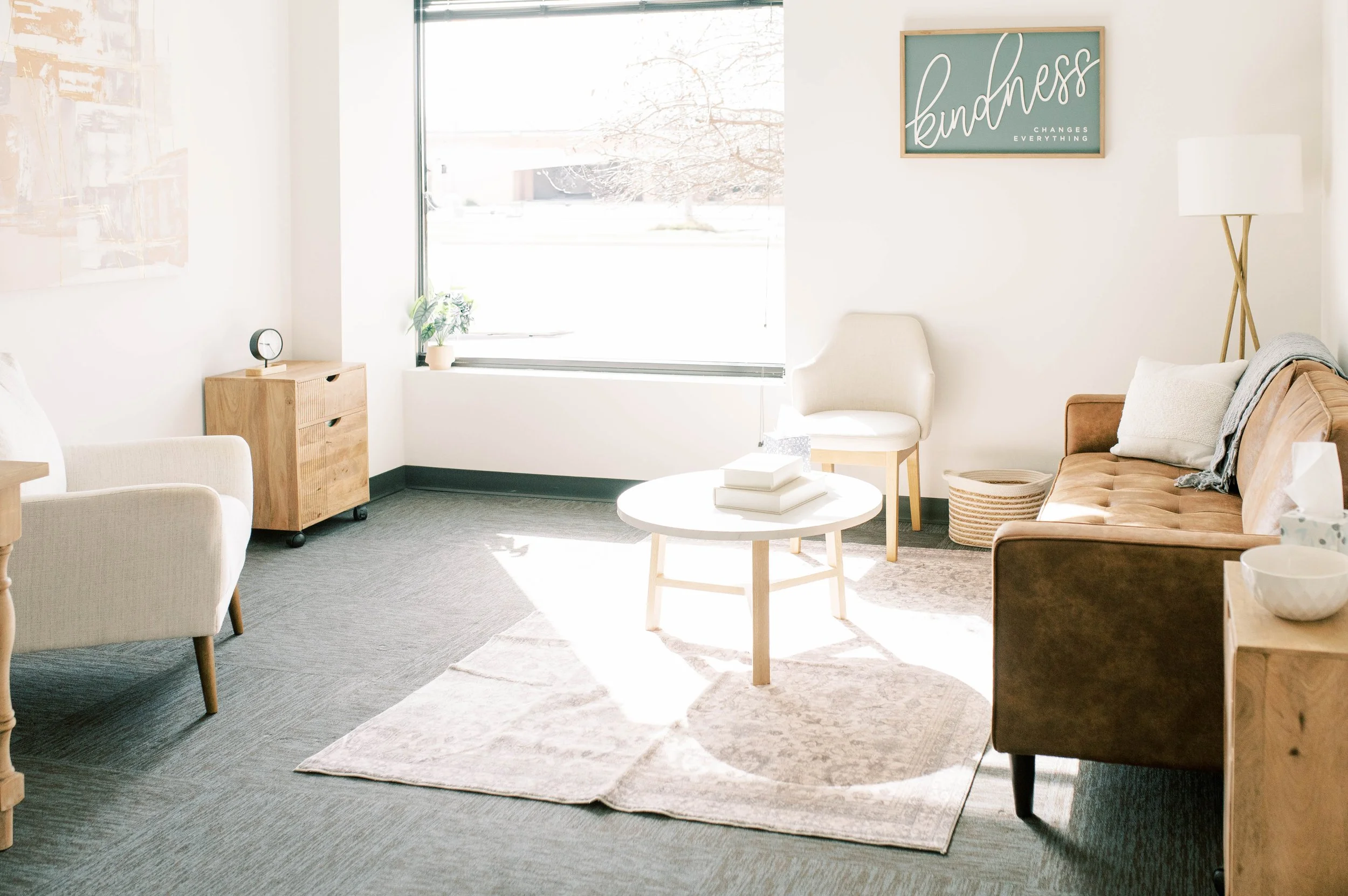 Bright living room with white walls, large window, and various furniture including a beige armchair, a brown sofa, a white round coffee table, a wooden side table, and a tall floor lamp. Decor includes a green sign with white script that says 'kindness changes everything' and a potted plant on the windowsill.