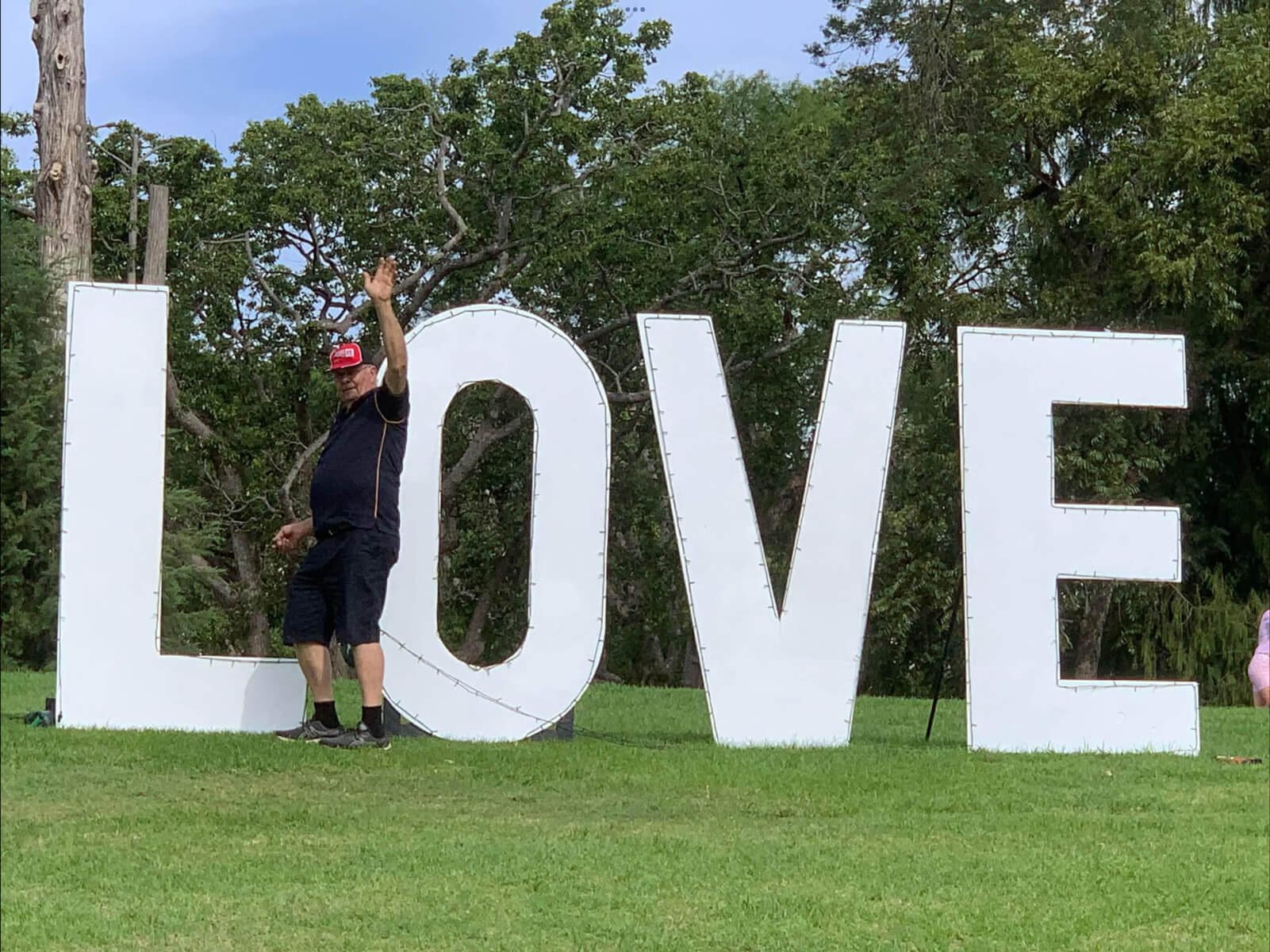 A man waving and standing next to large white letters spelling "LOVE" on a grassy field with trees in the background.