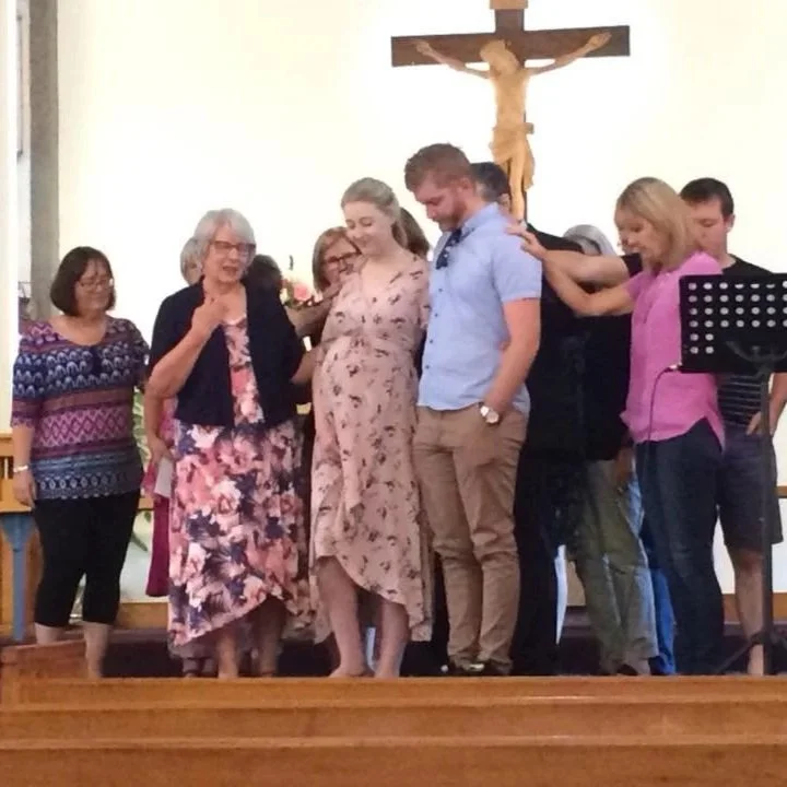 Group of people, including a pregnant woman, praying in a church with a crucifix hanging on the wall behind them.