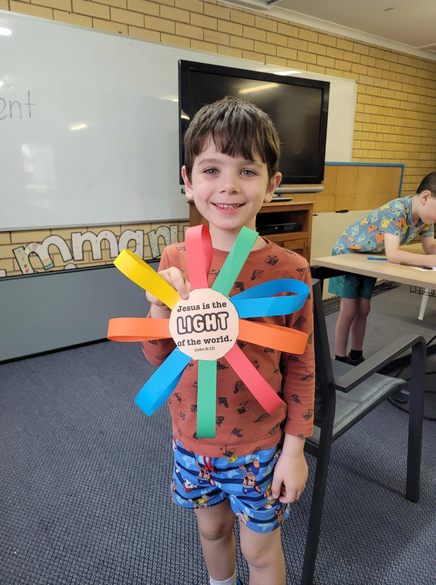 A smiling young boy holding colorful paper ribbons with a round sign that says, 'Jesus is the light of the world' in a classroom setting, with another boy working at a table in the background.