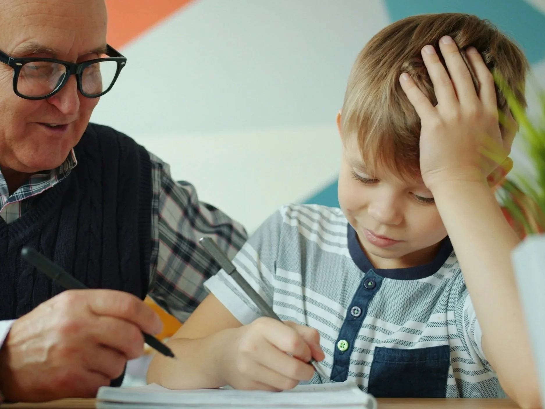 An elderly man wearing glasses and a checkered sweater helps a young boy with his schoolwork at a desk, holding a pen and pointing to paper.
