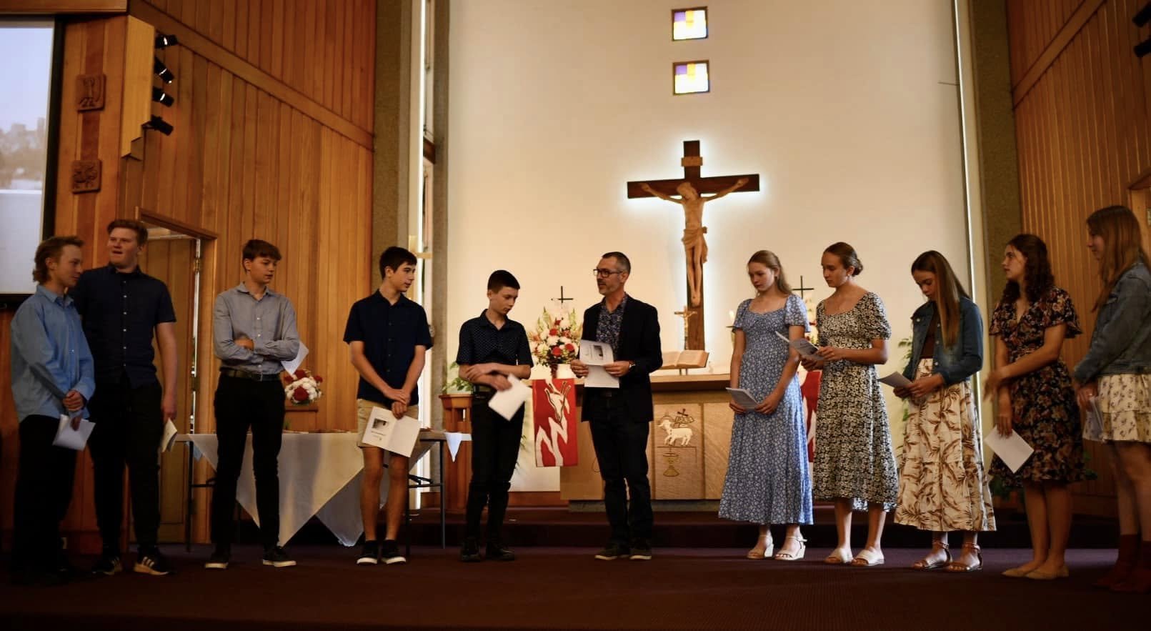 A group of young people stands in a church inside, receiving awards from a man in black. The church has wooden walls, a large crucifix behind the group, and stained glass windows.