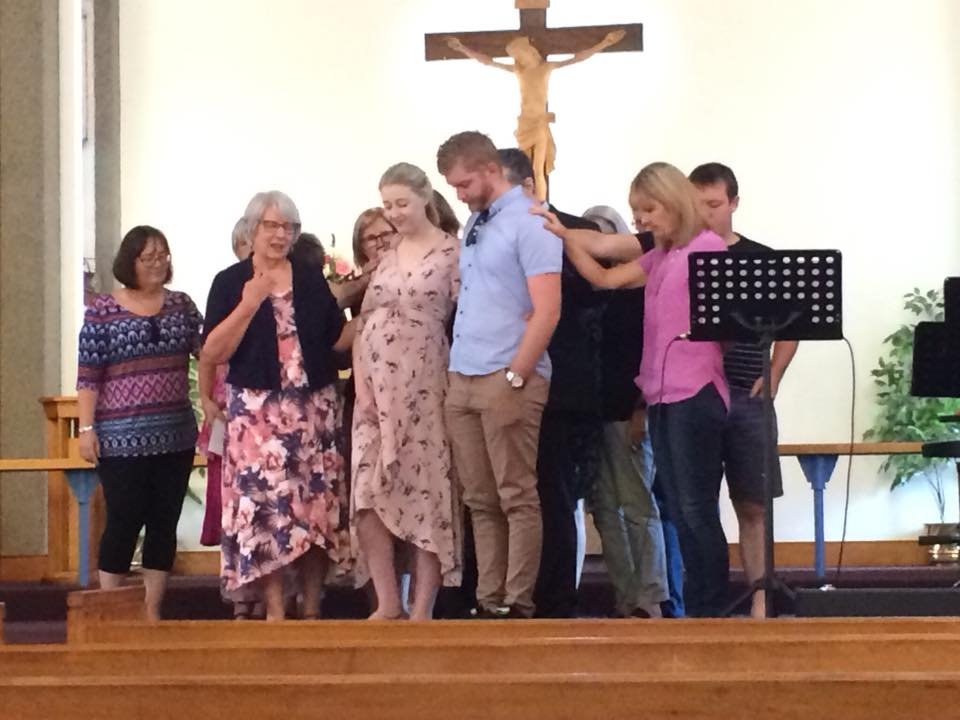 A group of people standing on a church altar, some holding or touching a pregnant woman, with a crucifix hanging above them.