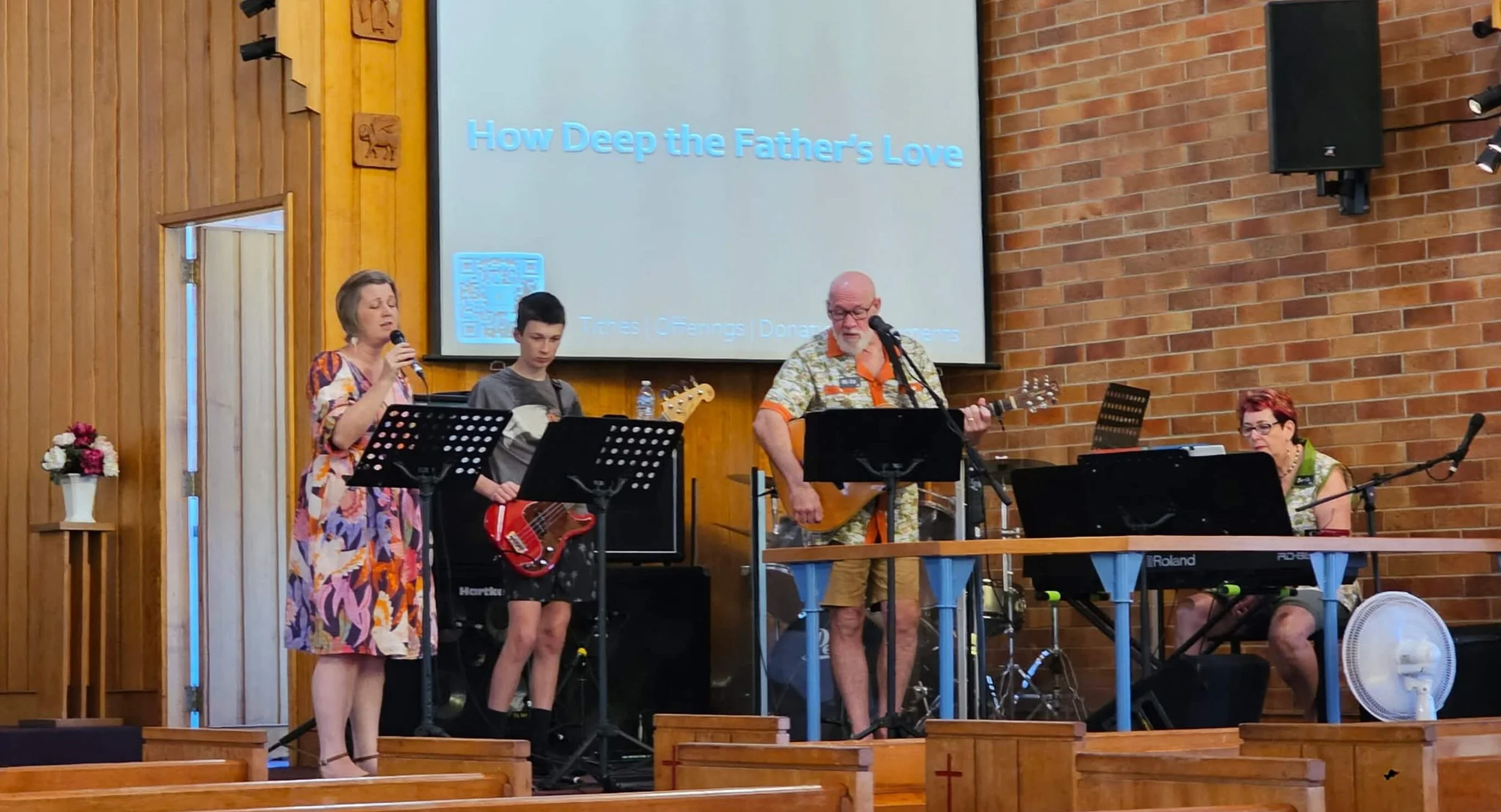 A group of four musicians performing on a stage inside a church with a brick wall back and a wooden wall side, and a projector screen displaying the words "How Deep the Father's Love."