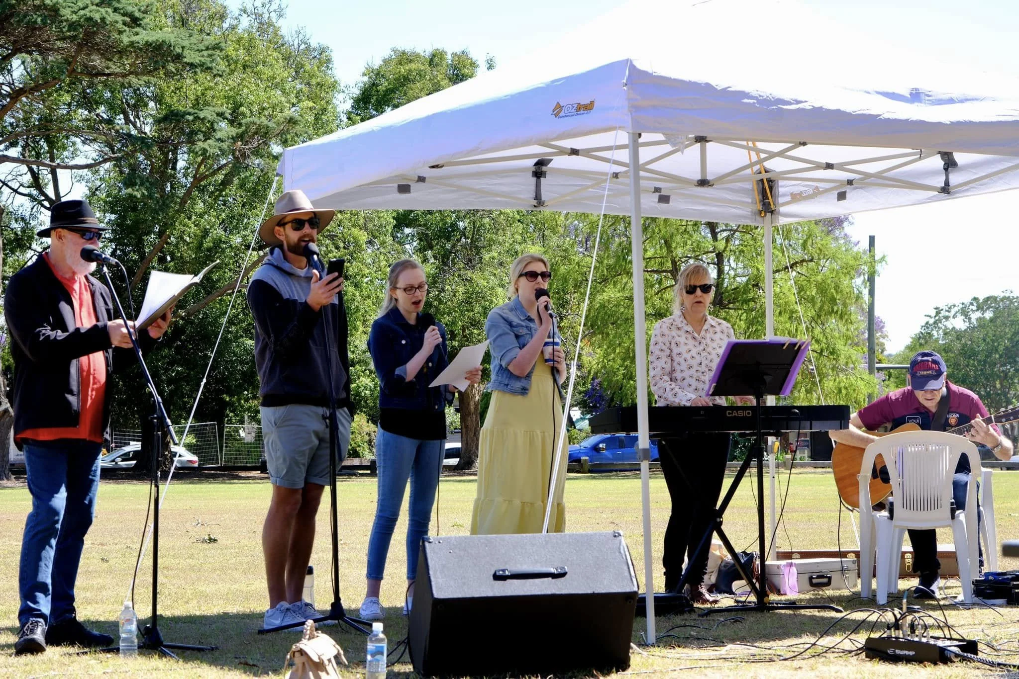 Group of people performing outdoor music show under a white canopy with trees in the background, some holding microphones and instruments.