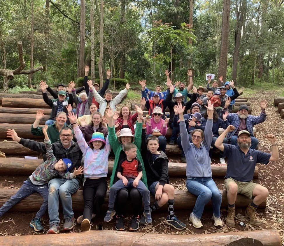 Group of people sitting on logs in a forest, raising their hands and smiling at the camera.