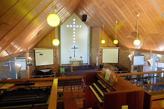 Interior of a church with a wooden ceiling, illuminated by hanging spherical lights, featuring an altar with a cross and two candles, and a piano in the foreground.
