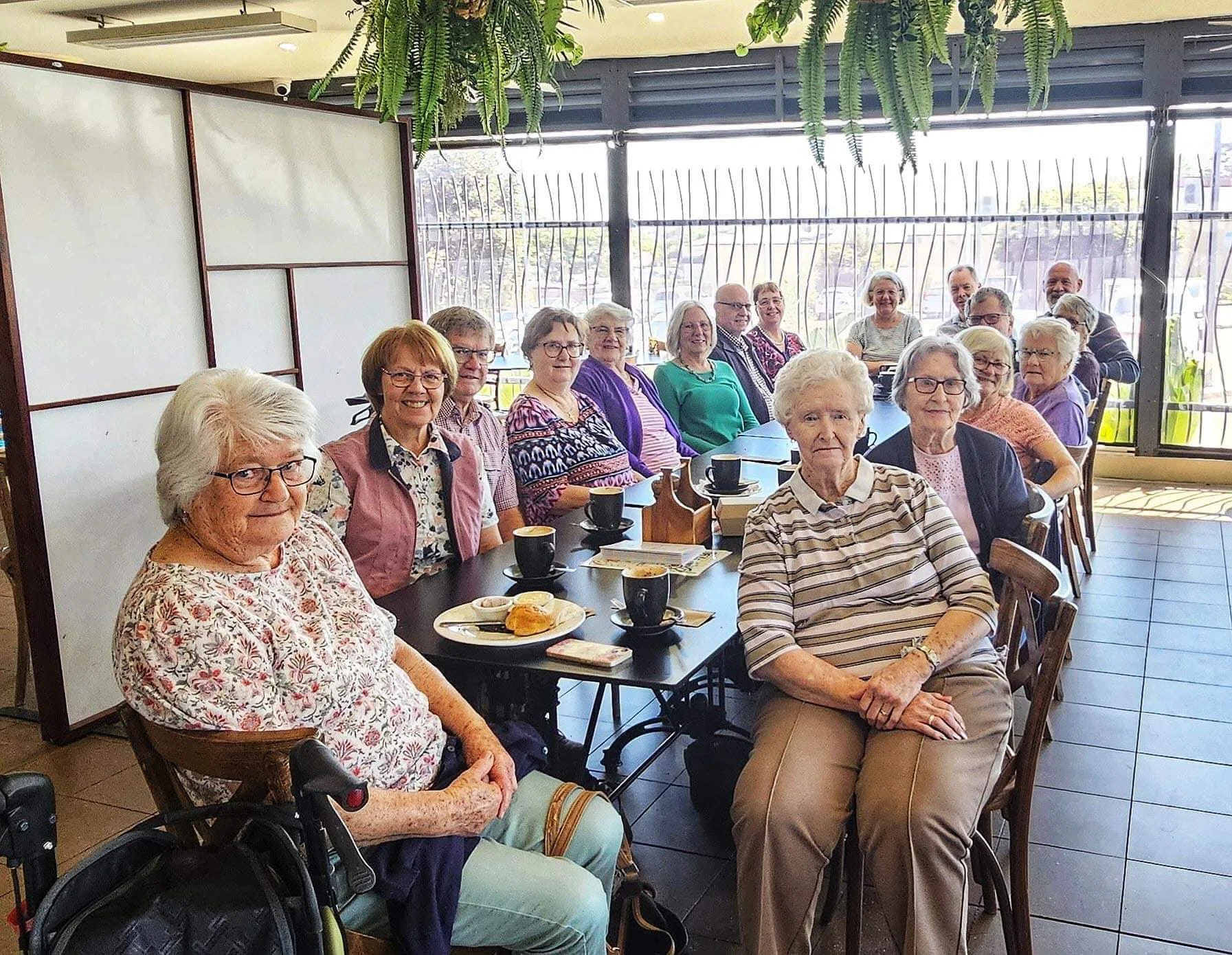 A group of elderly people, mostly women, sitting around a long table in a bright cafe or restaurant, enjoying coffee and breakfast. The setting includes large windows, hanging plants, and a decorative partition.